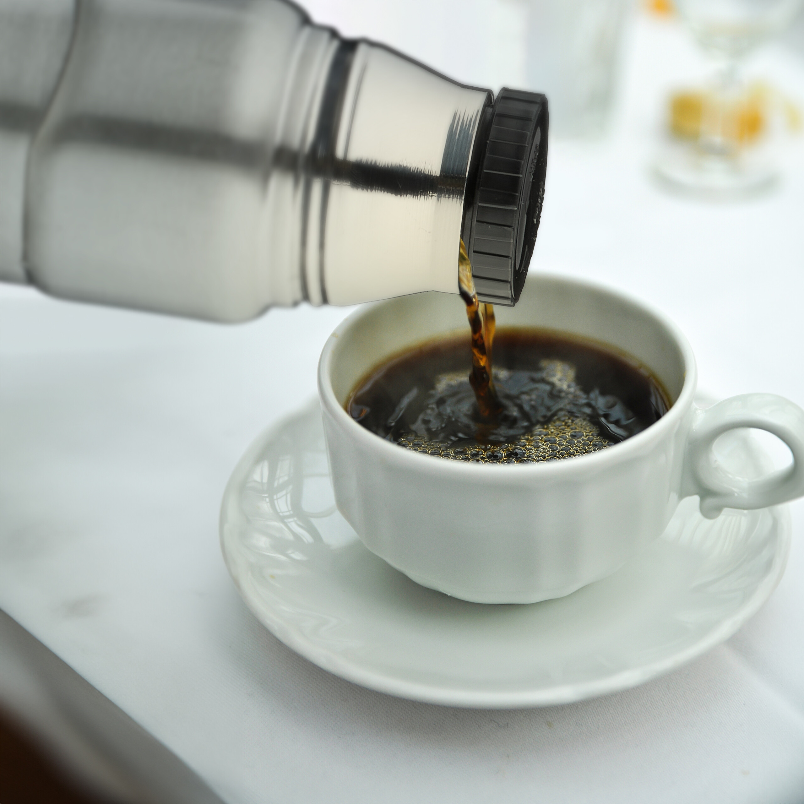 Eine Thermoskanne gießt dunklen Kaffee in eine weiße Tasse auf einer Untertasse.