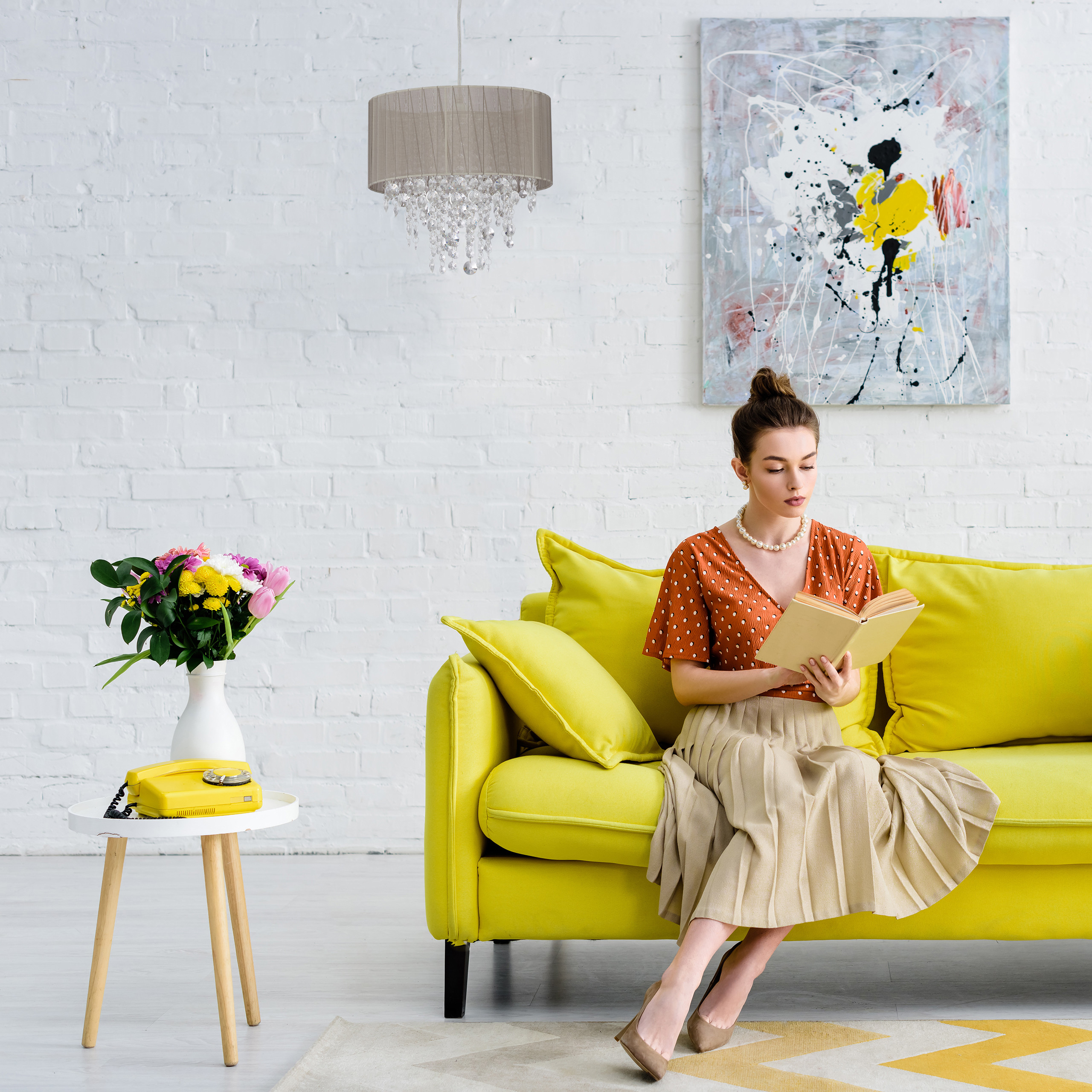 Eine Frau in einem orangen Oberteil liest auf einem gelben Sofa ein Buch in einem hellen Zimmer mit bunter Kunst an der Wand.