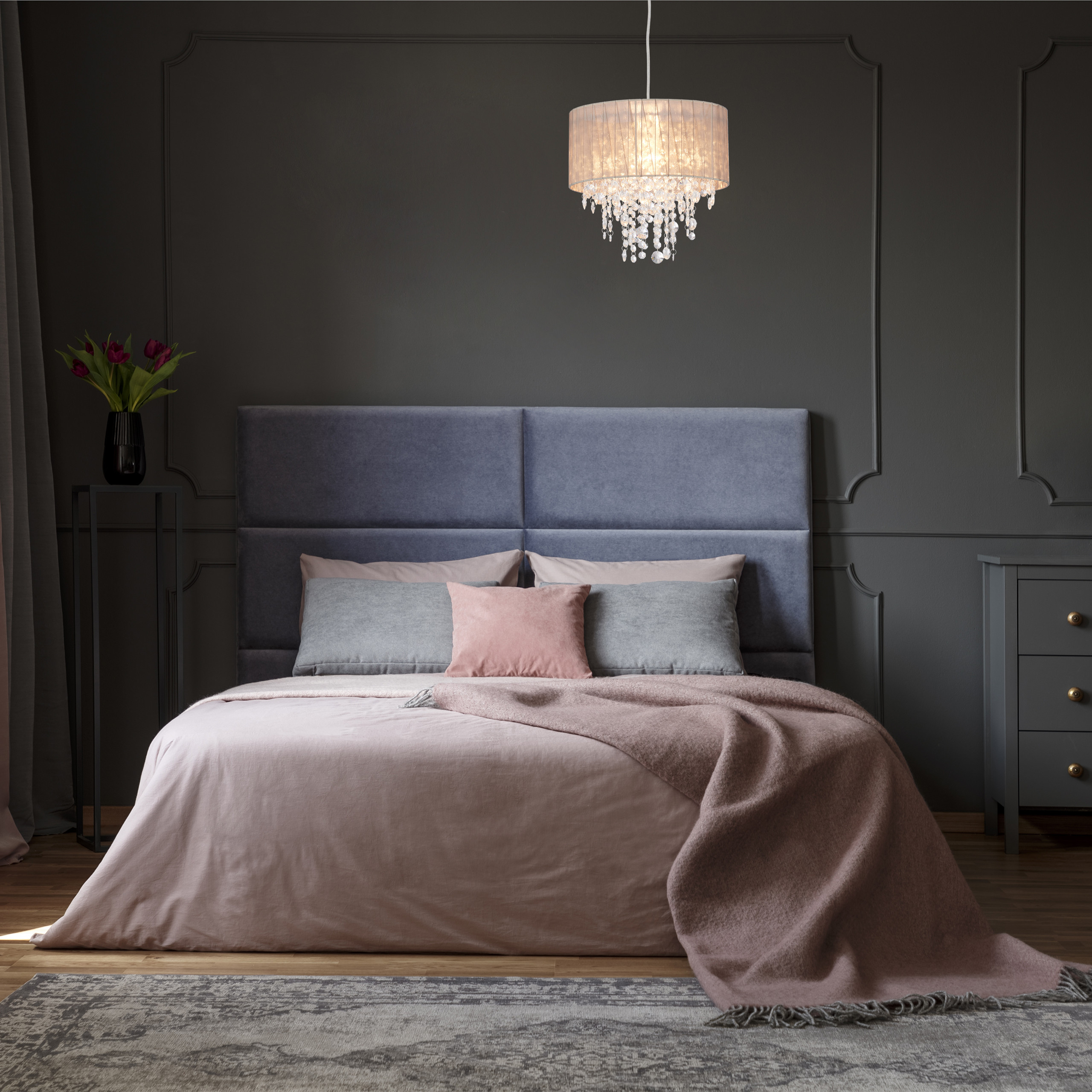 Ein Schlafzimmer mit blauem Kopfteil, rosa Bettwäsche, grauen Kissen, Kronleuchter und dunkelgrauen Wandpaneelen.