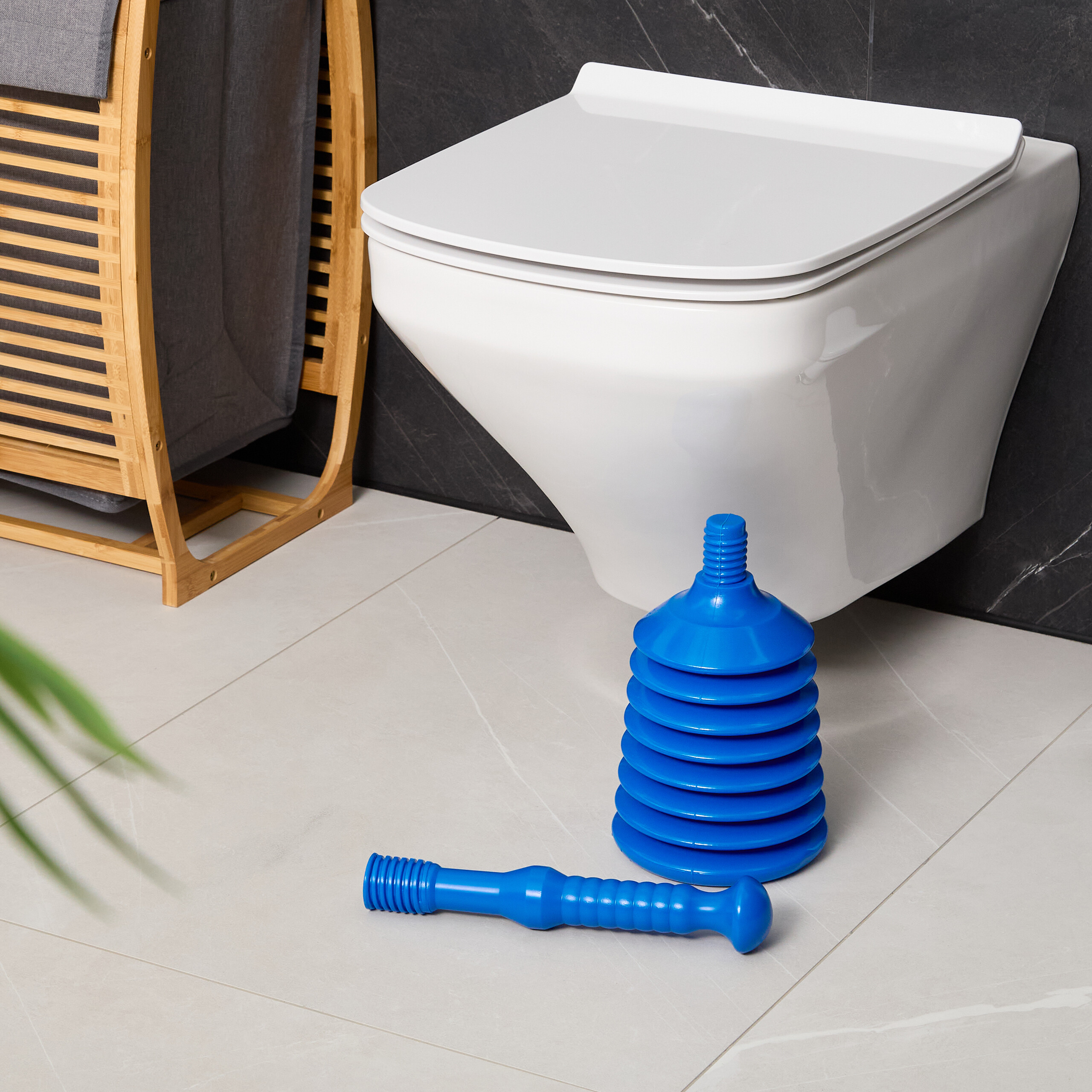 Ein blauer, zerlegter Pümpel liegt auf weißen Fliesen vor einer Toilette und einem Holzwäschekorb.