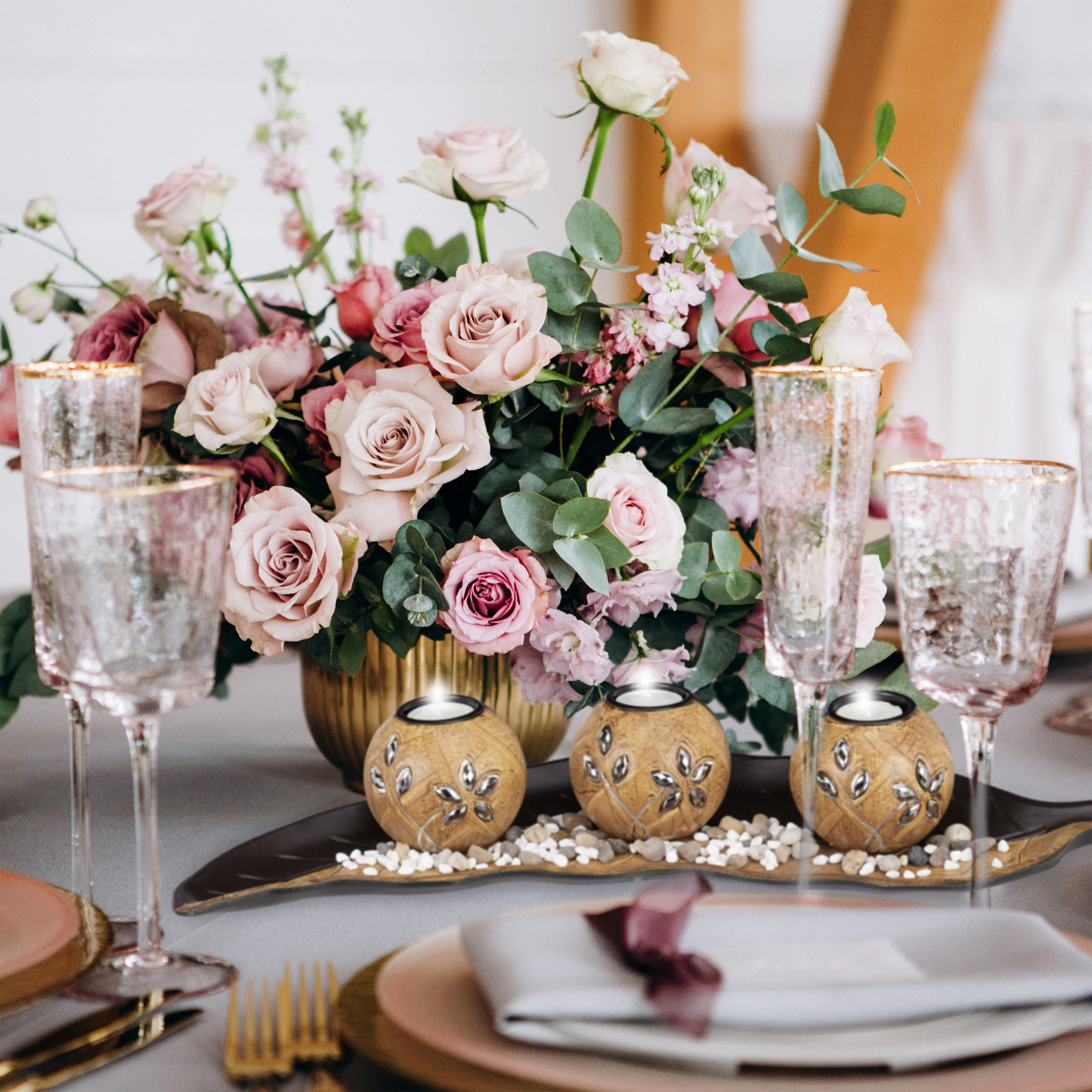 Tischdekoration mit großem rosa Blumenarrangement, drei kleinen Goldkerzen auf Tablett und Kristallgläsern.
