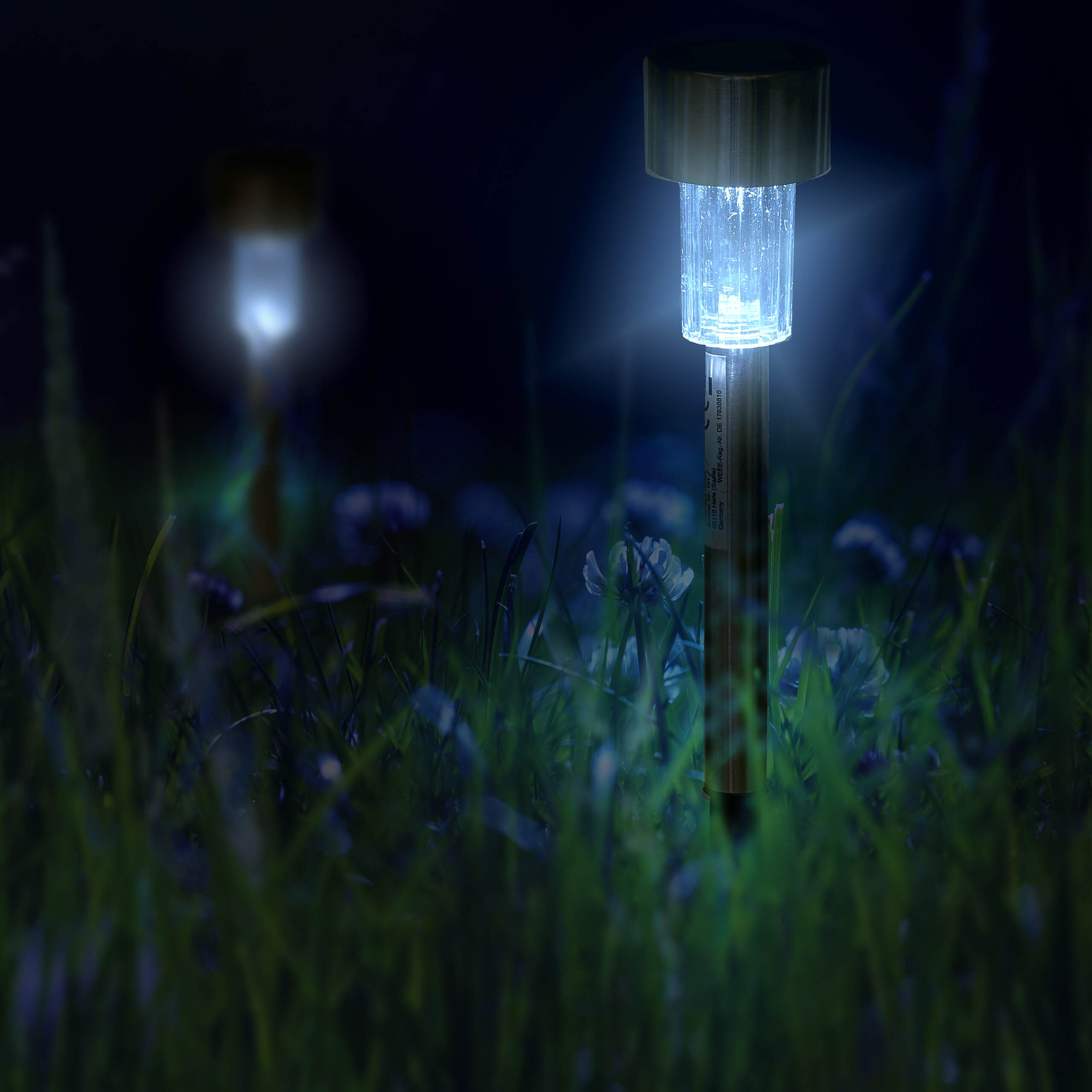 Eine blau leuchtende Solarlampe steht im dunklen, hohen Gras, im Hintergrund ist eine zweite Lampe verschwommen zu sehen.