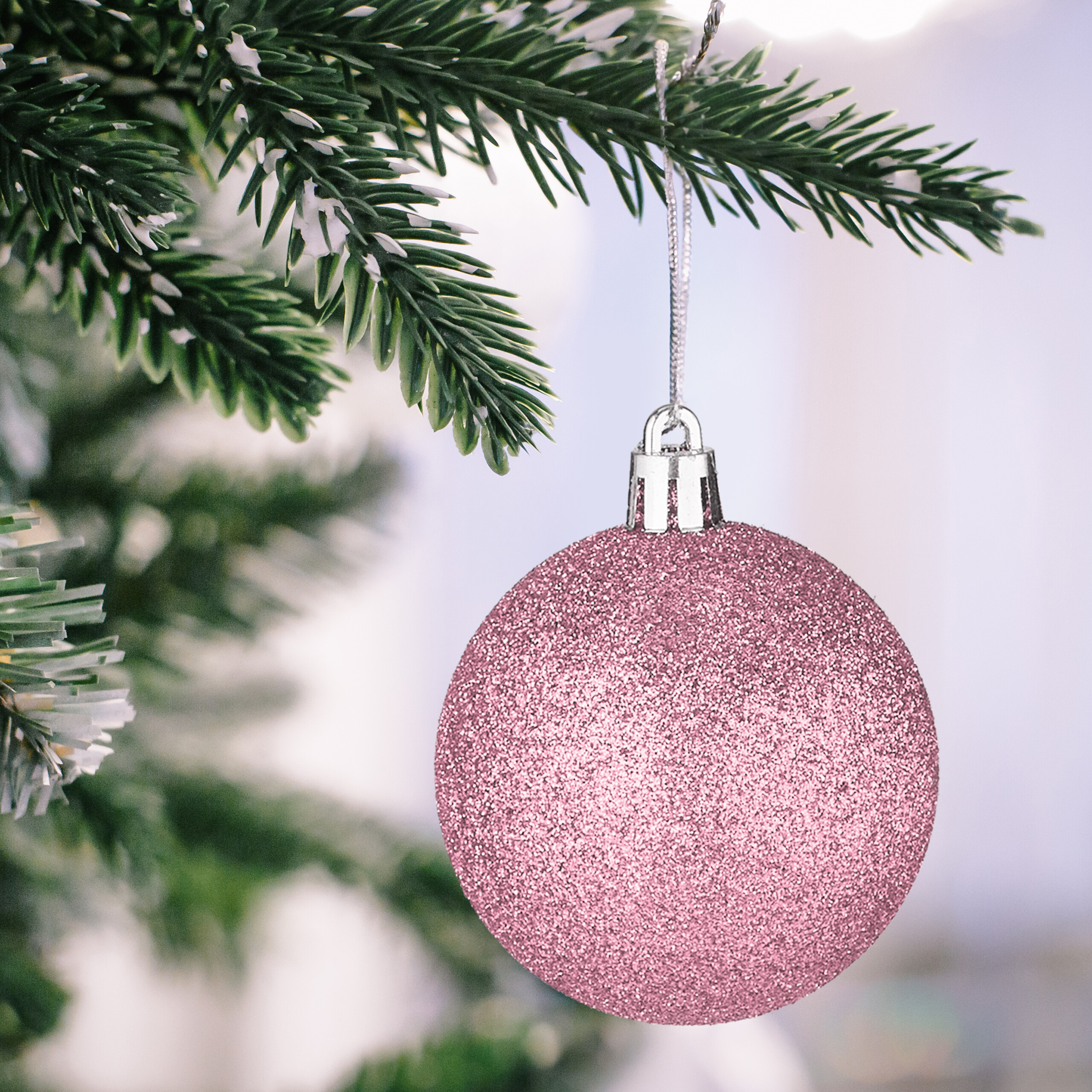 Eine glitzernde rosa Weihnachtskugel hängt an einem verschneiten Tannenzweig.