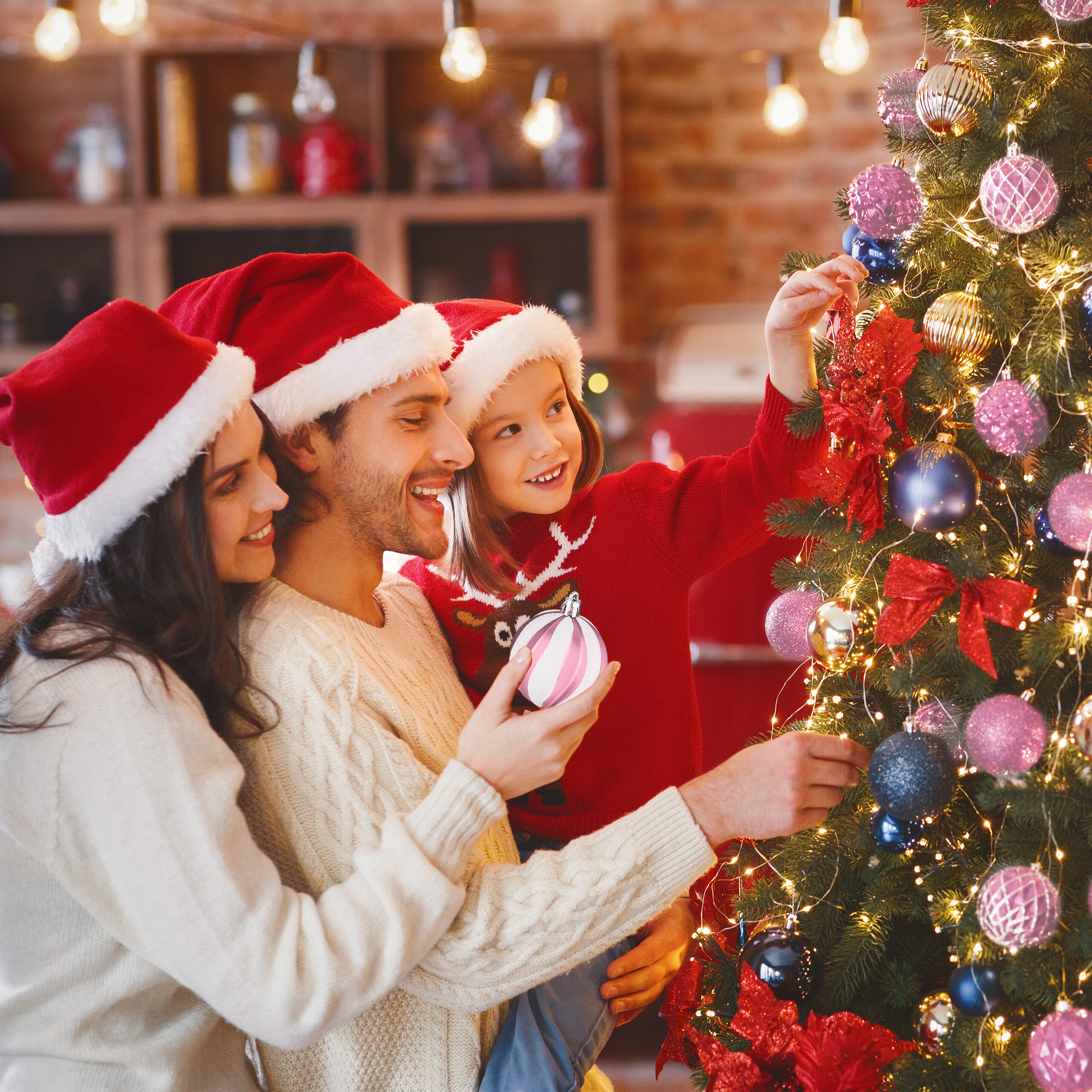Eine Familie mit Weihnachtsmützen schmückt gemeinsam den Weihnachtsbaum.
