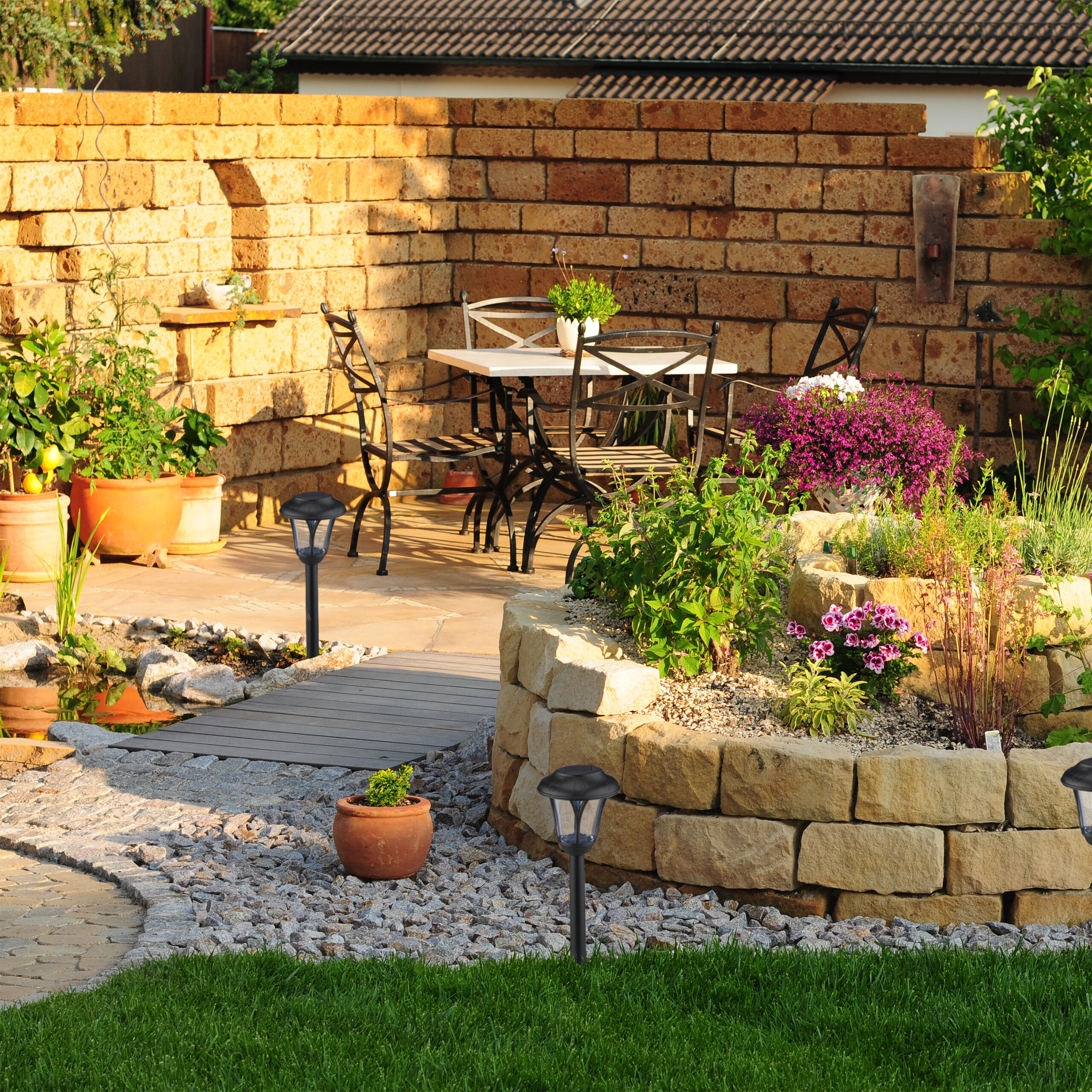 Ein sonniger Garten mit Steinmauer, Hochbeet, Teich, Terrassentisch und Solarlichtern entlang eines Kieswegs.