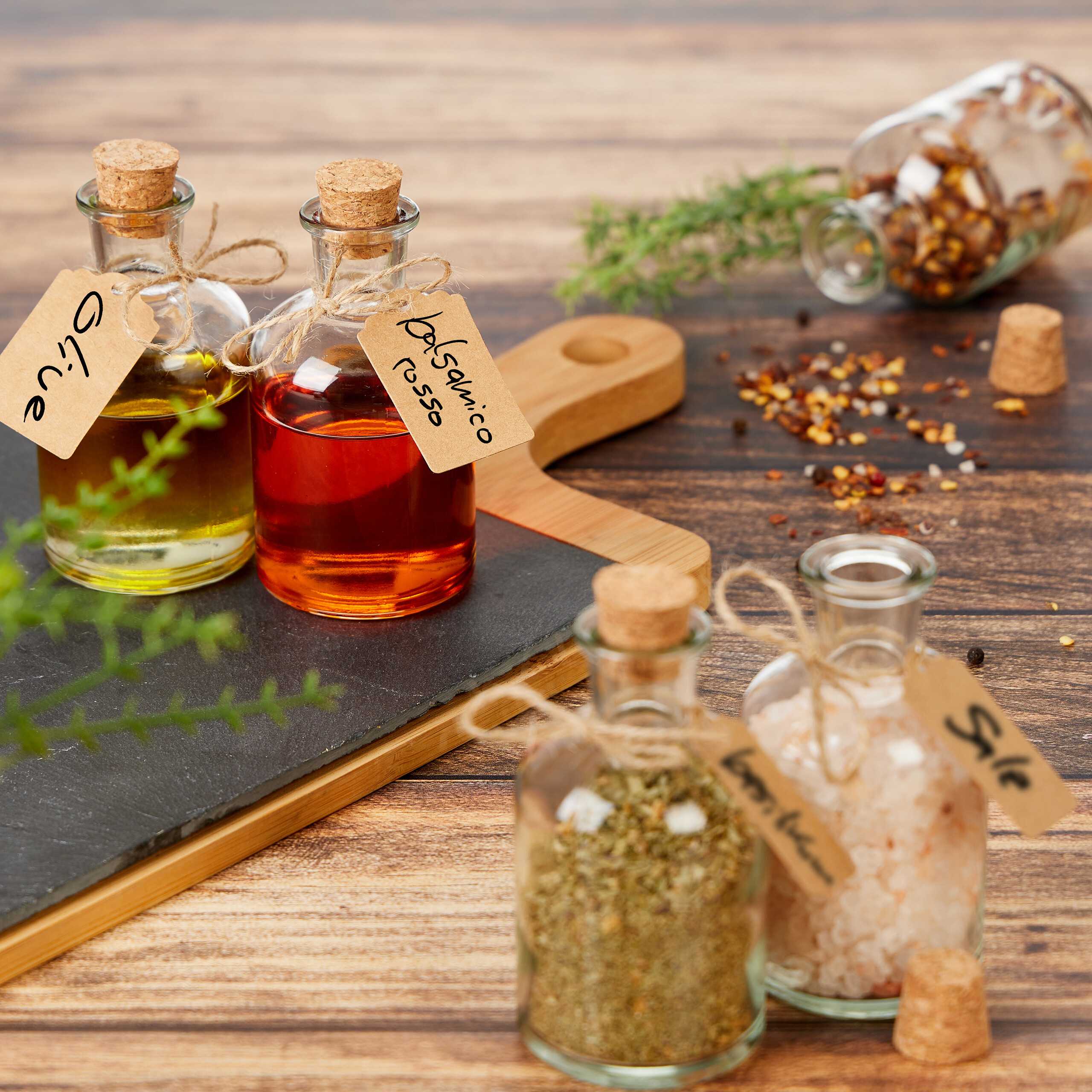 Glasflaschen mit Öl, Essig, Kräutern und Salz auf einem Holzbrett.