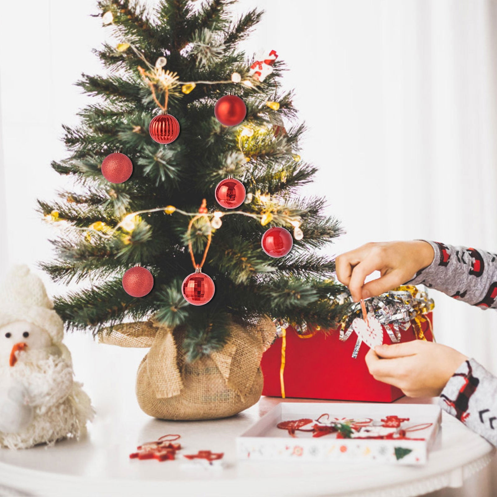 Eine Person schmückt einen kleinen Weihnachtsbaum mit roten Kugeln, Lichtern und einer Schneemannfigur auf dem Tisch.