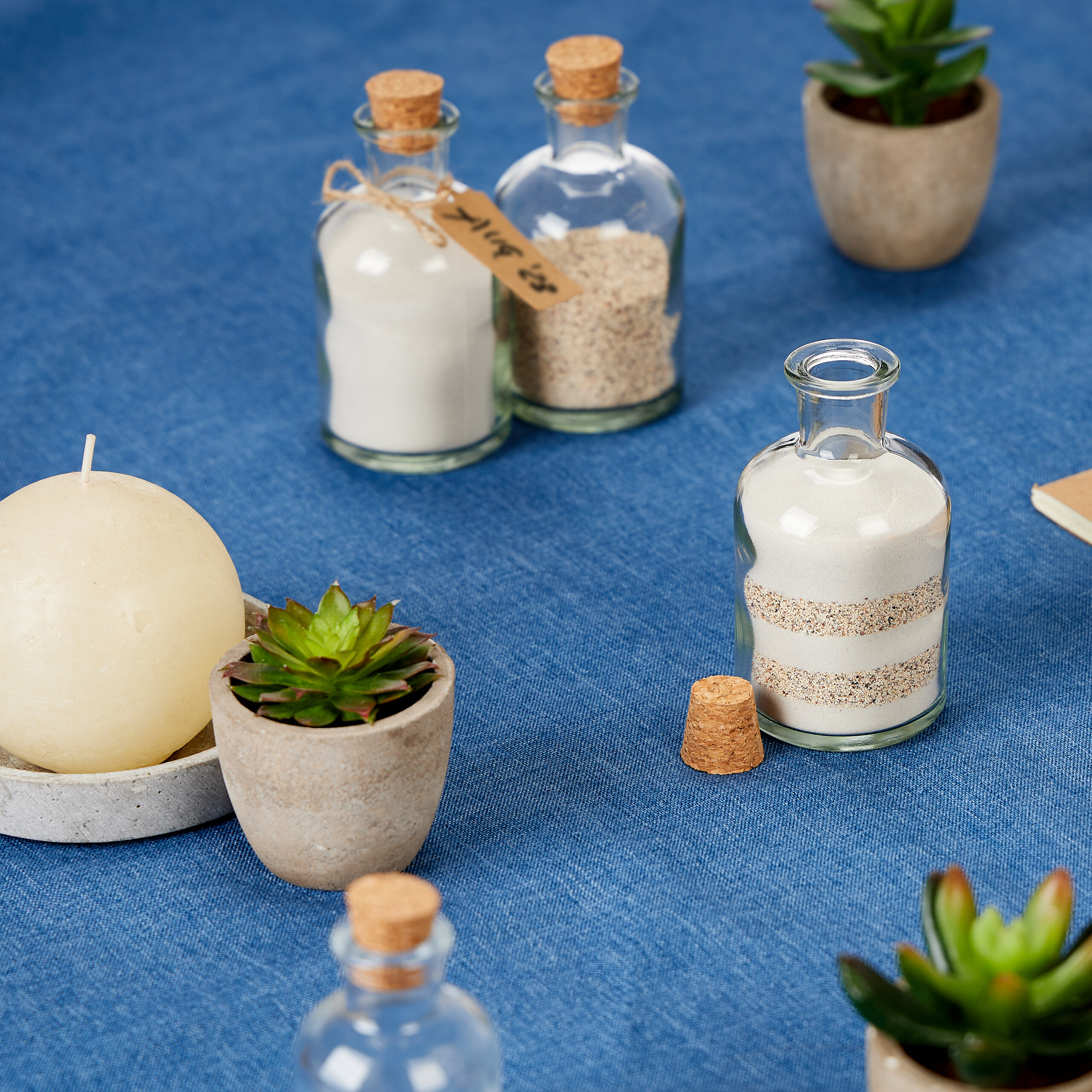 Glasflaschen mit Sand, Sukkulenten und eine Kerze auf blauem Untergrund.
