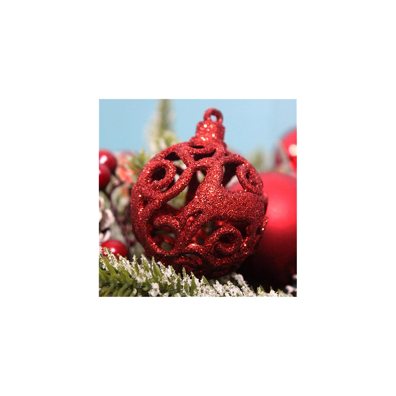 Eine rote, glitzernde Weihnachtskugel mit ausgestanztem Muster liegt auf verschneiten Tannenzweigen.