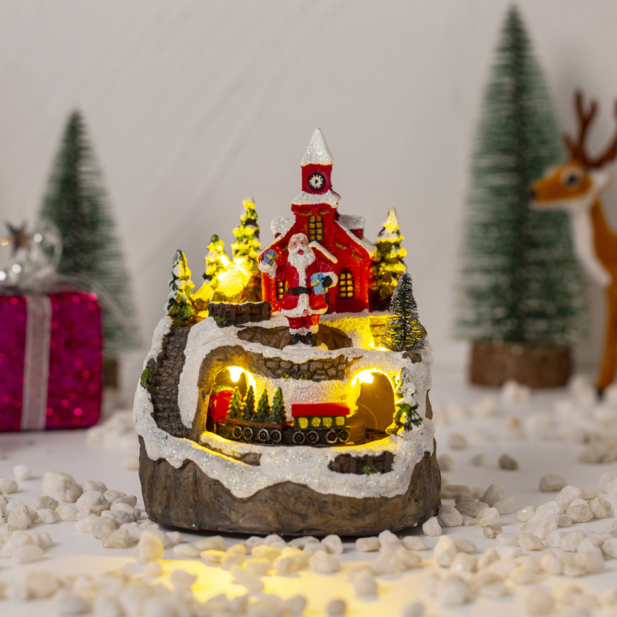 Beleuchtete Weihnachtsdorf-Figur mit Santa, Kirche, Zug und Kunstschnee, umgeben von Bäumen und Geschenken.