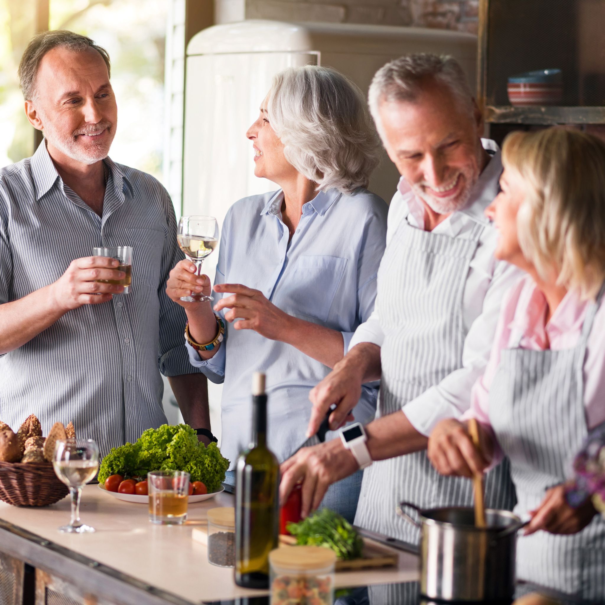 Vier ältere Freunde kochen zusammen in einer Küche, trinken Wein und lächeln.