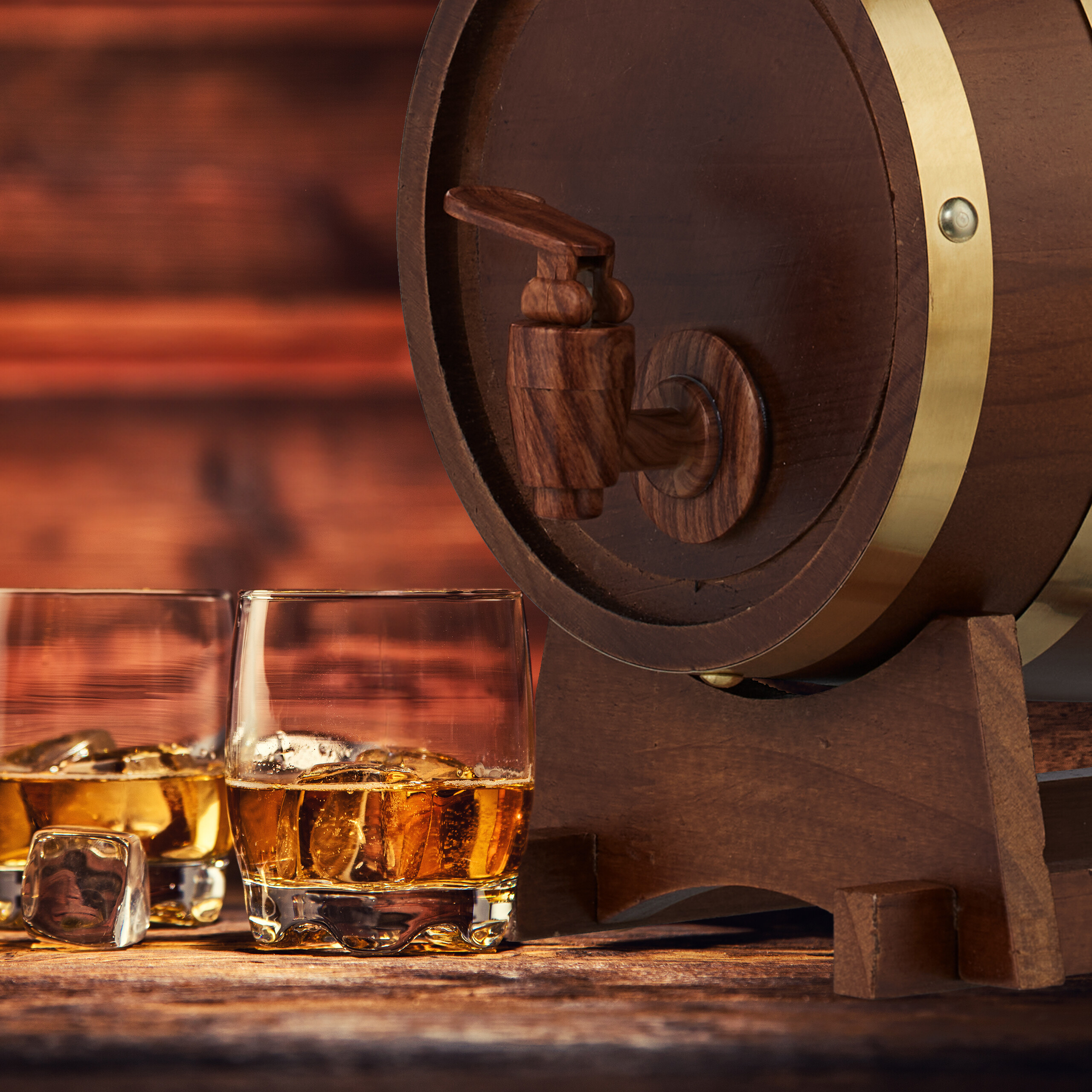 Ein Holzfass mit Zapfhahn und zwei Gläser Whiskey mit Eis auf einem Holztisch.