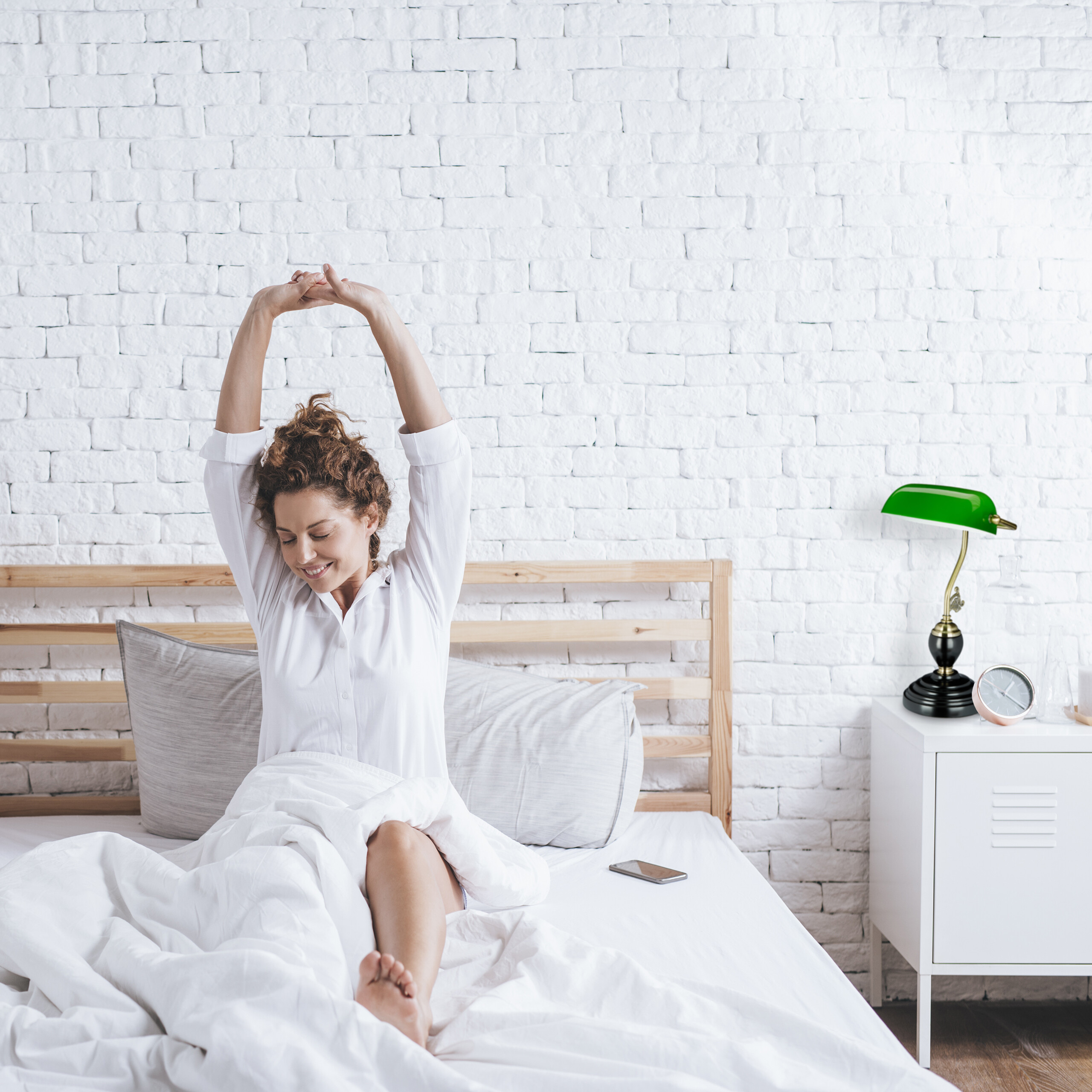 Eine Frau im weißen Hemd streckt sich im Bett vor einer weißen Backsteinwand mit grüner Lampe.