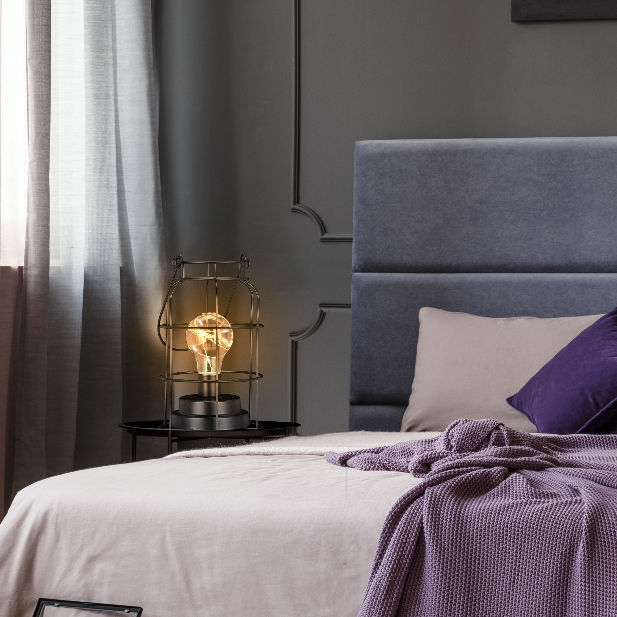Moderne Lampe mit Glühbirne auf Nachttisch neben gemütlichem Bett mit lilafarbenen Kissen.