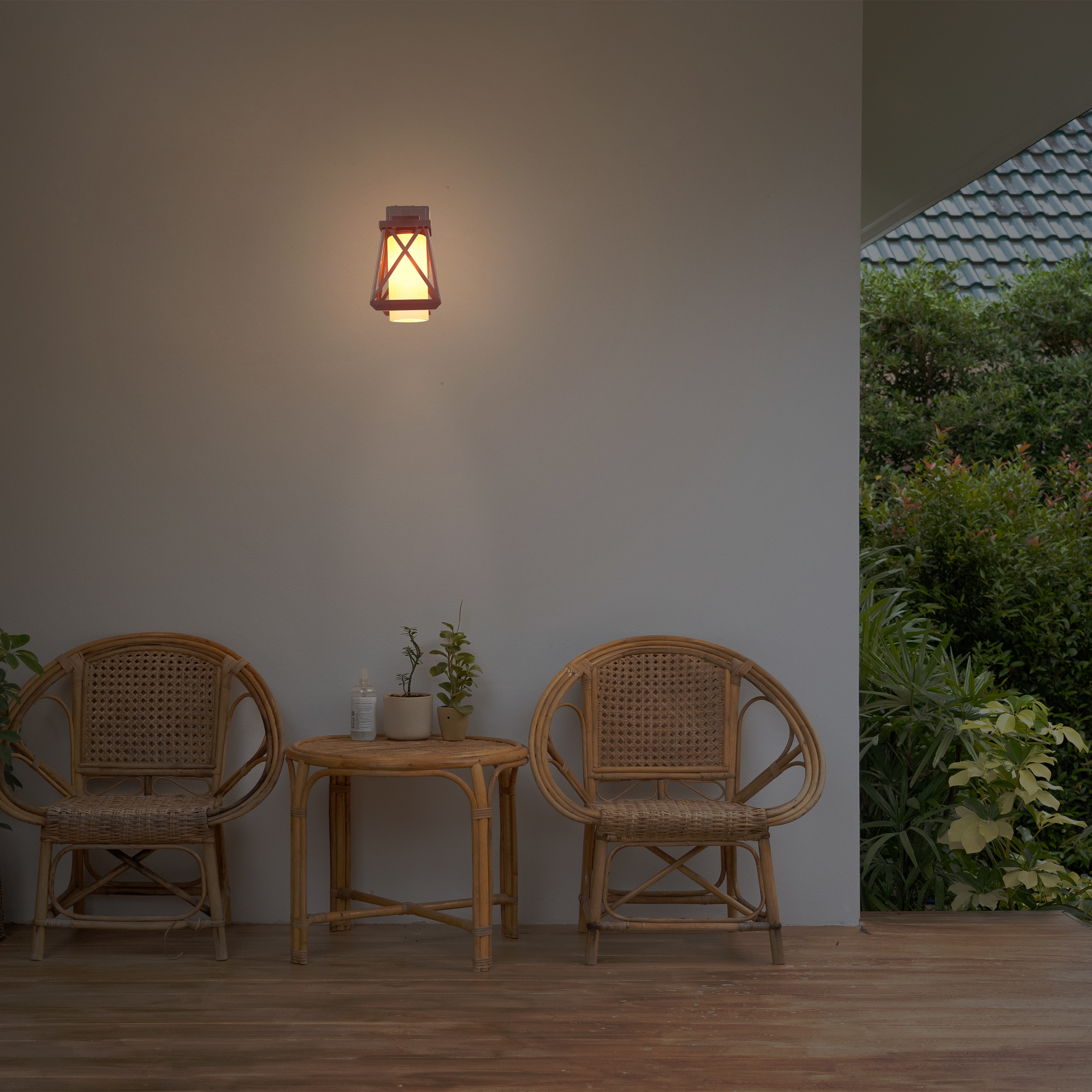 Eine Außenterrasse mit zwei Rattanstühlen, einem kleinen Tisch mit Topfpflanzen und einer leuchtenden Wandlampe.