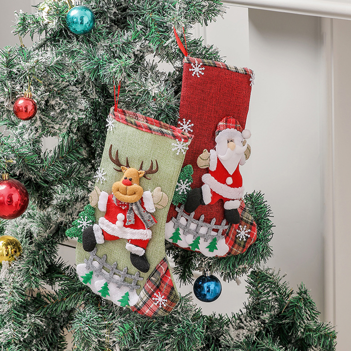 Zwei Weihnachtssocken, eine grüne mit Rentier und eine rote mit Weihnachtsmann, hängen an einem verschneiten Baum.