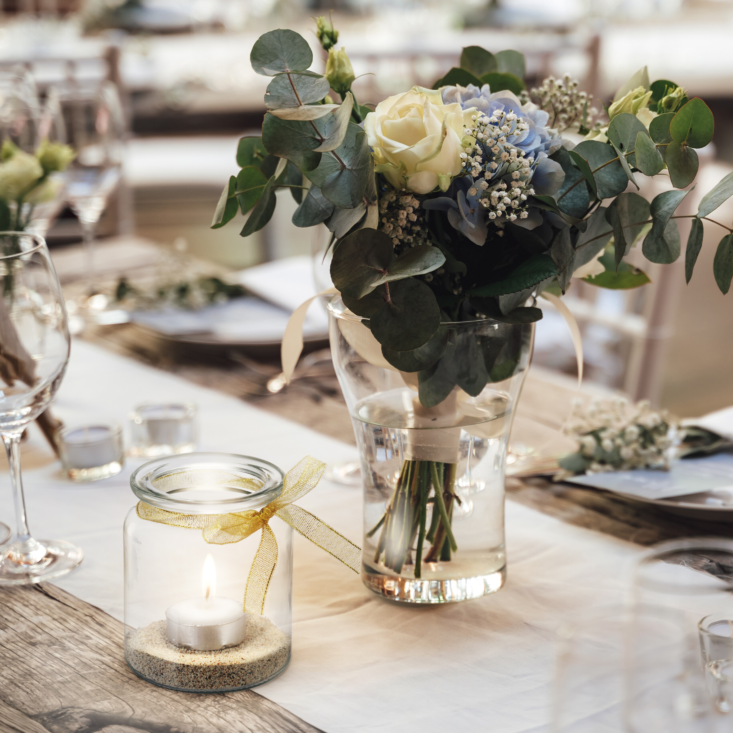 Eine Hochzeitstischdeko mit Vase, weißen Rosen und Eukalyptus neben einer brennenden Kerze im Glas mit Sand.
