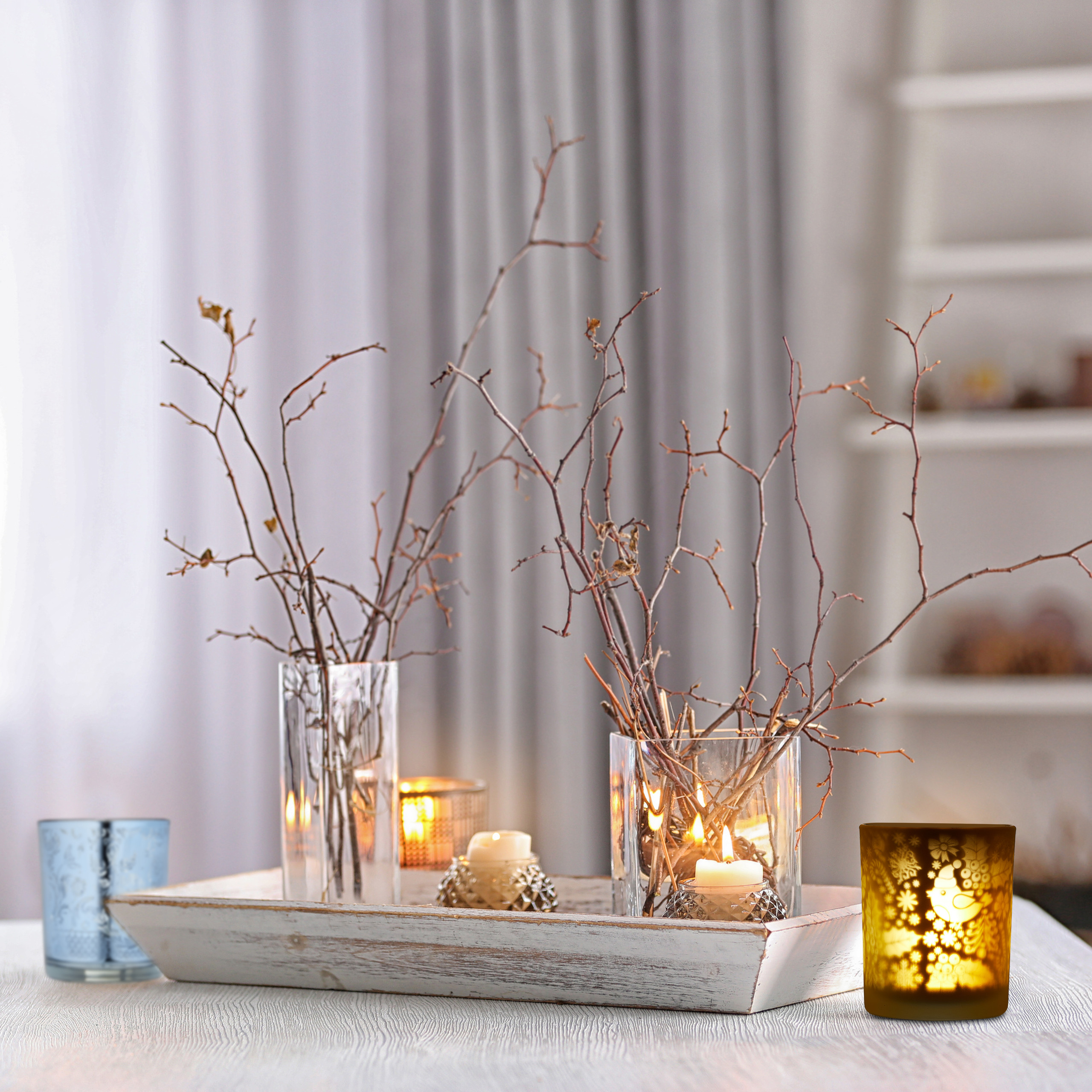Ein Holztablett mit Glasvasen, trockenen Zweigen und brennenden Kerzen steht auf einem Tisch.