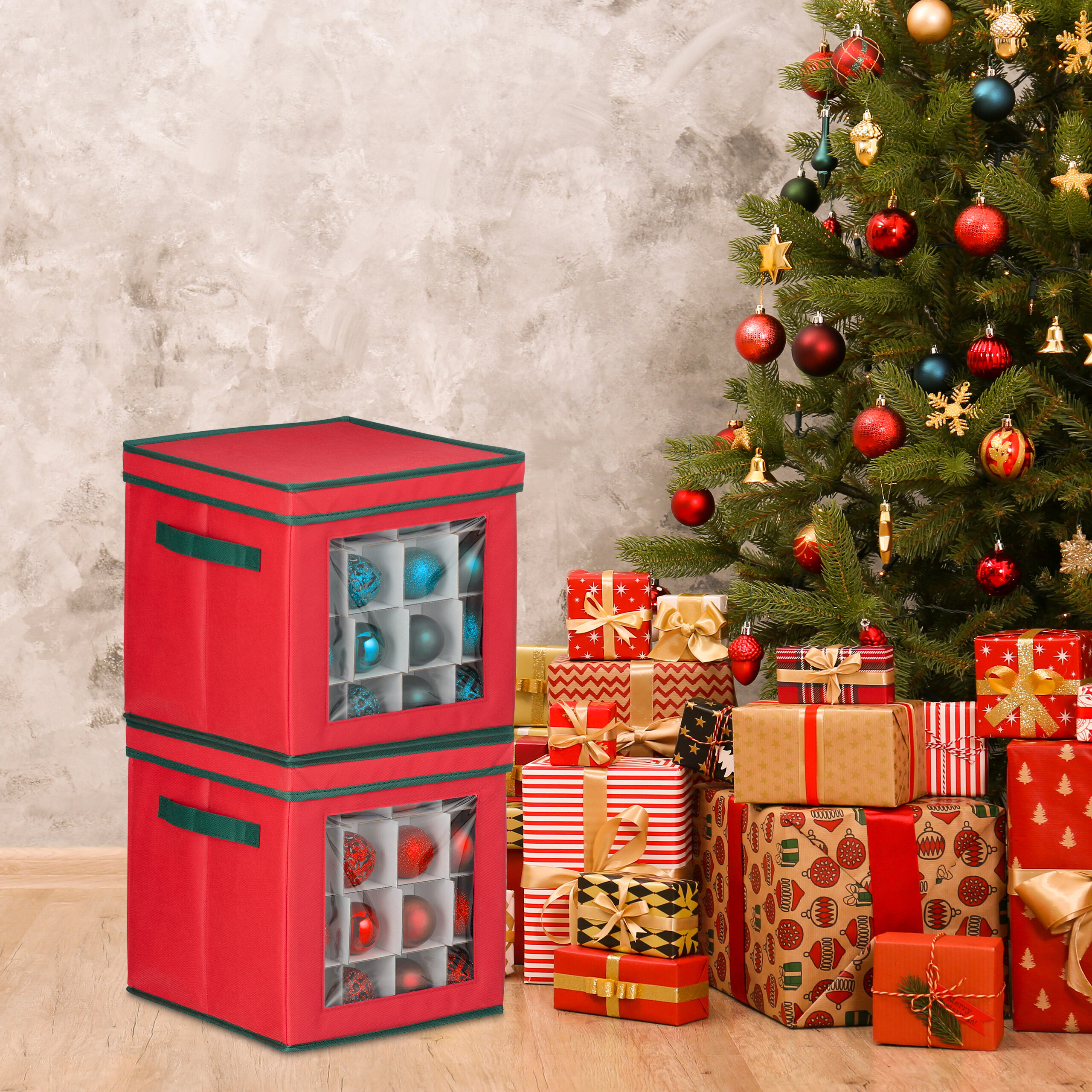 Zwei rote Aufbewahrungsboxen mit Weihnachtskugeln stehen vor einer beleuchteten Tanne mit Geschenken.