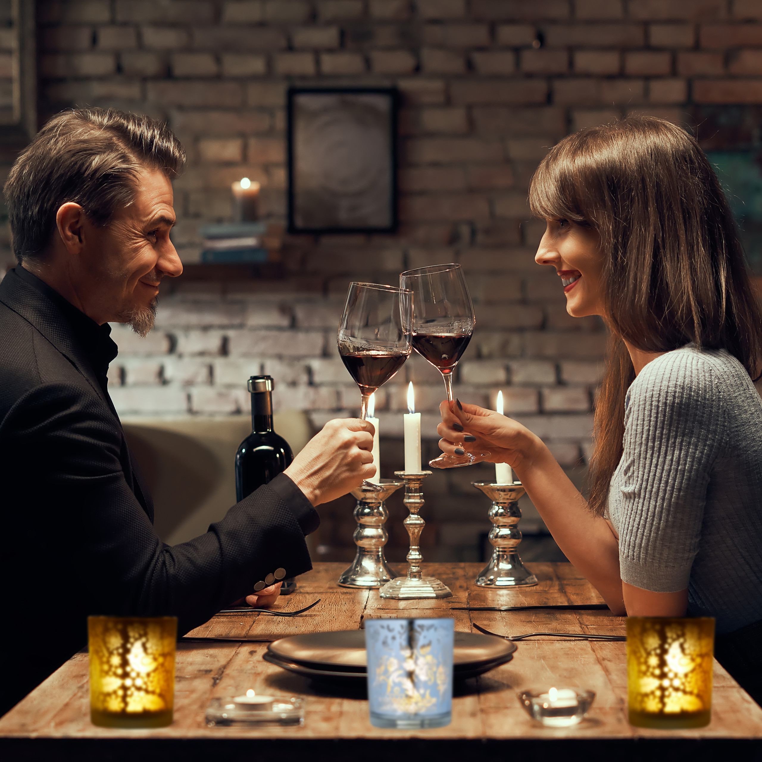 Ein Paar sitzt an einem Holztisch bei Kerzenschein und stößt mit Rotwein an.