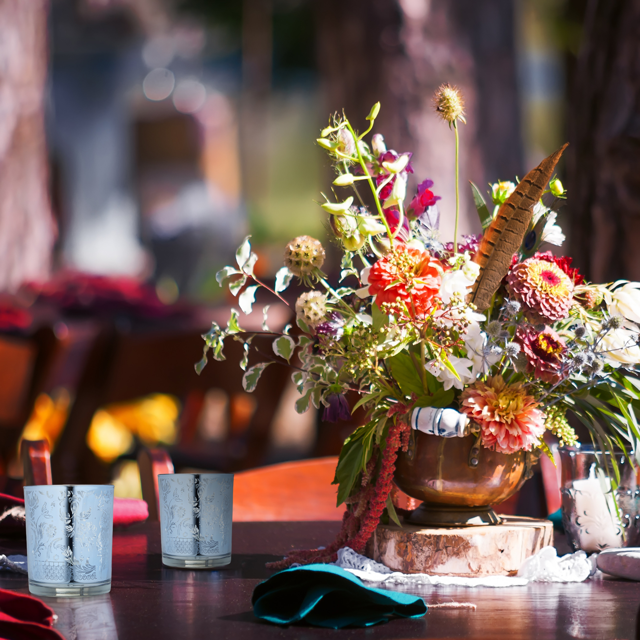Ein hölzerner Tisch mit einer bunten Blumenvase und verzierten Gläsern vor einer verschwommenen Naturkulisse.