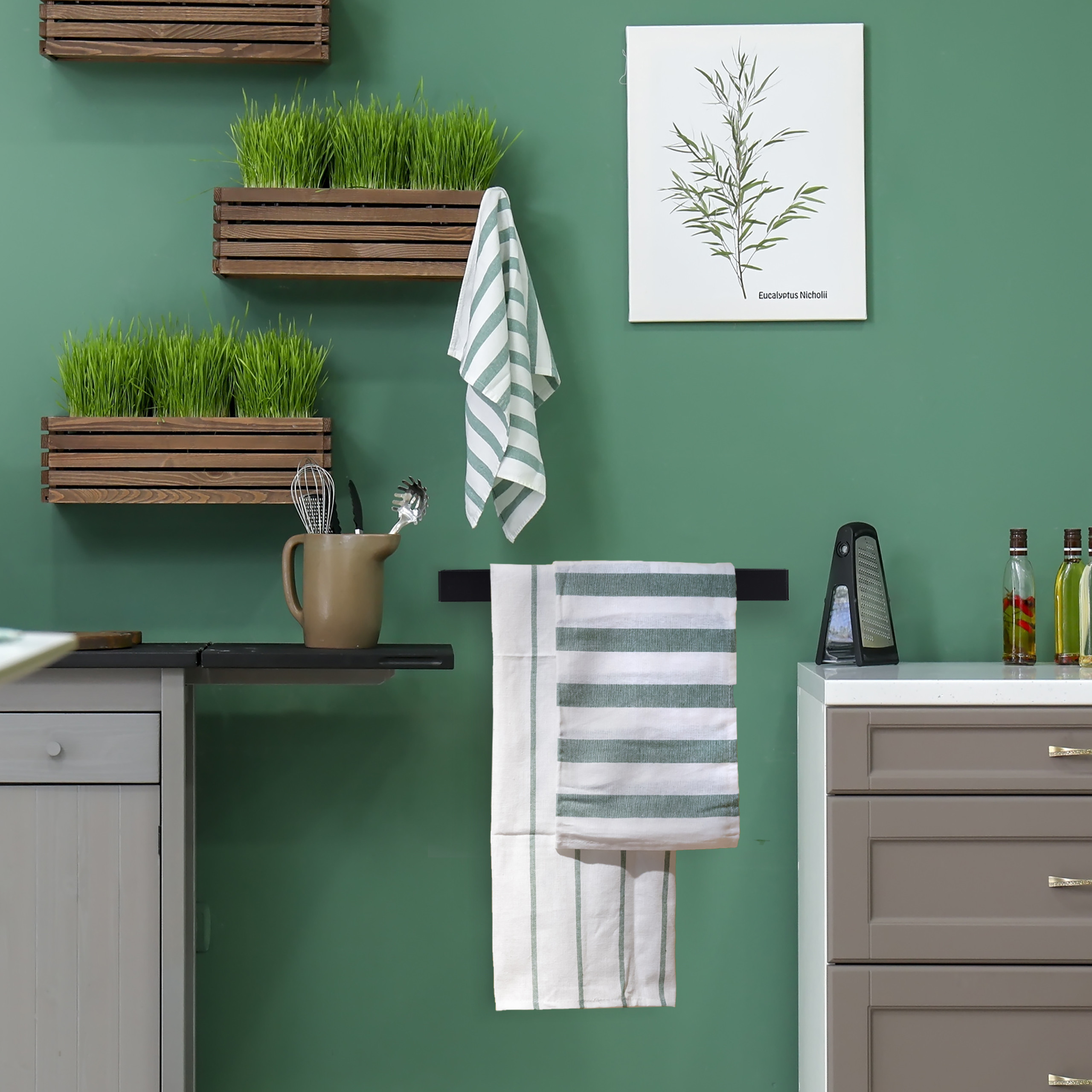 Grüne Wand mit Holzkisten voller Gras, hängenden Handtüchern, Küchenutensilien und einer Pflanzengrafik.