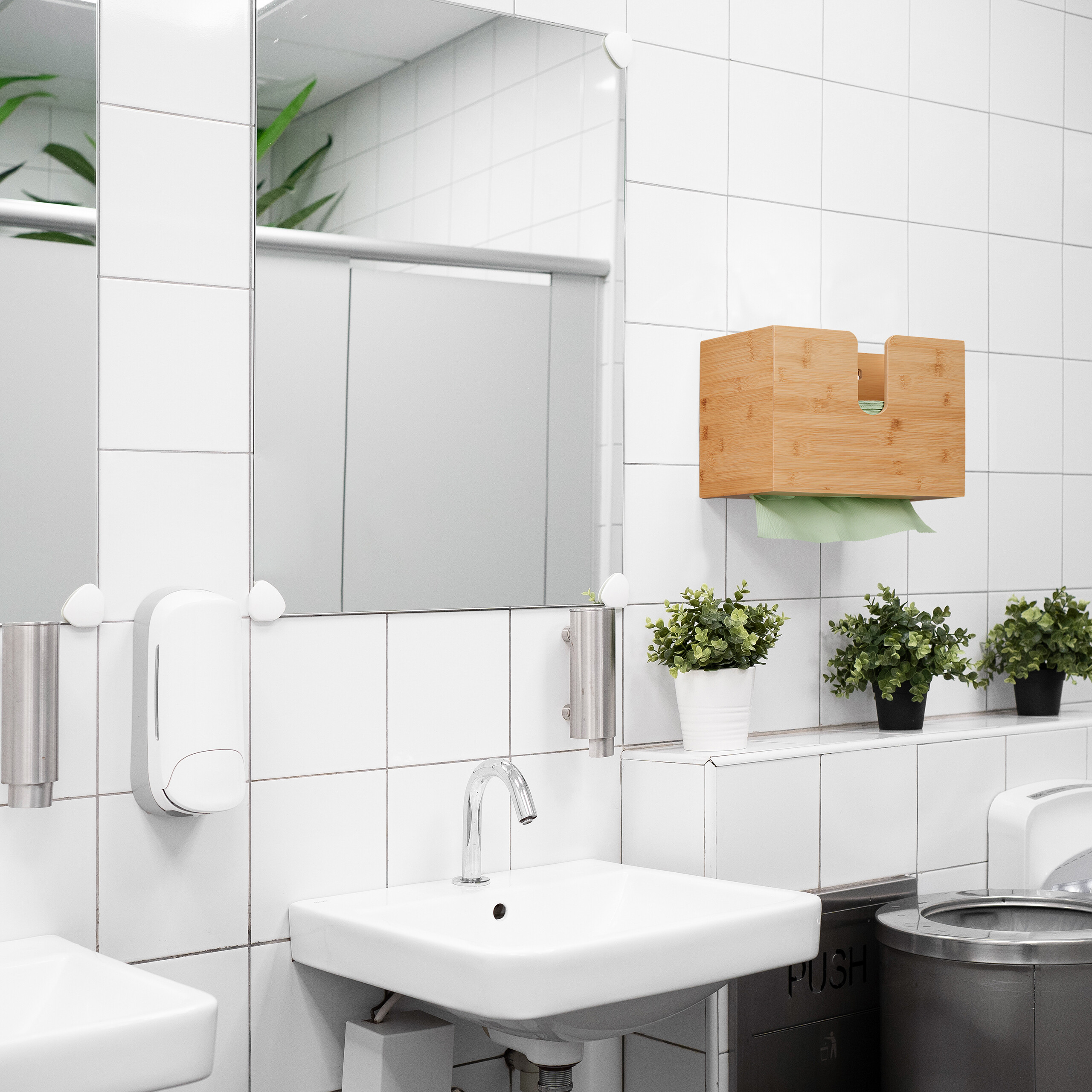 Ein weißes Waschbecken mit Spiegel, Seifenspender und Holztuchspender an einer weiß gefliesten Wand.