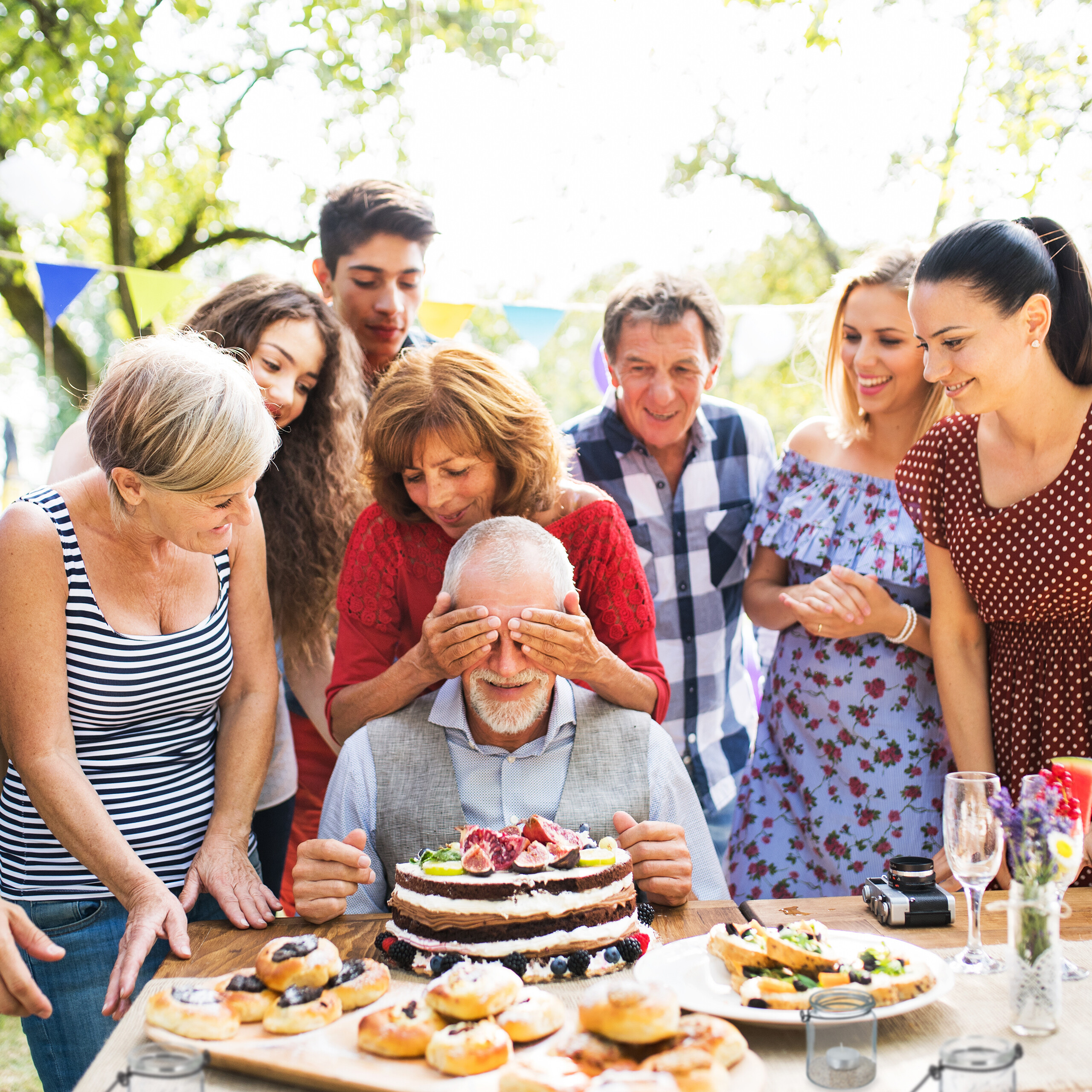Eine Familie versammelt sich um einen glücklichen älteren Mann, dem die Augen zugehalten werden, vor einer Geburtstagstorte.