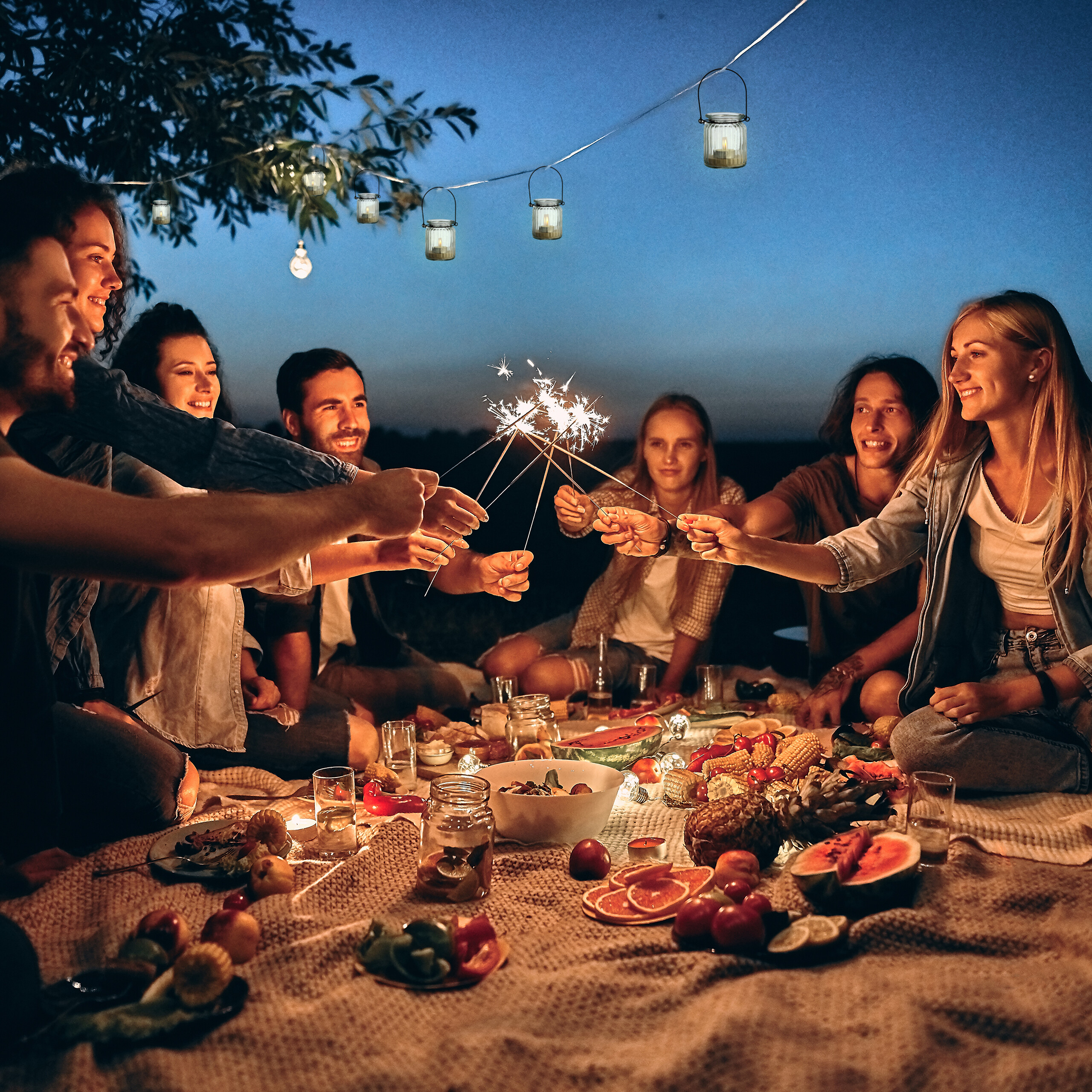 Freunde feiern draußen im Dunkeln ein Picknick, halten Wunderkerzen und sitzen auf einer Decke mit Essen.