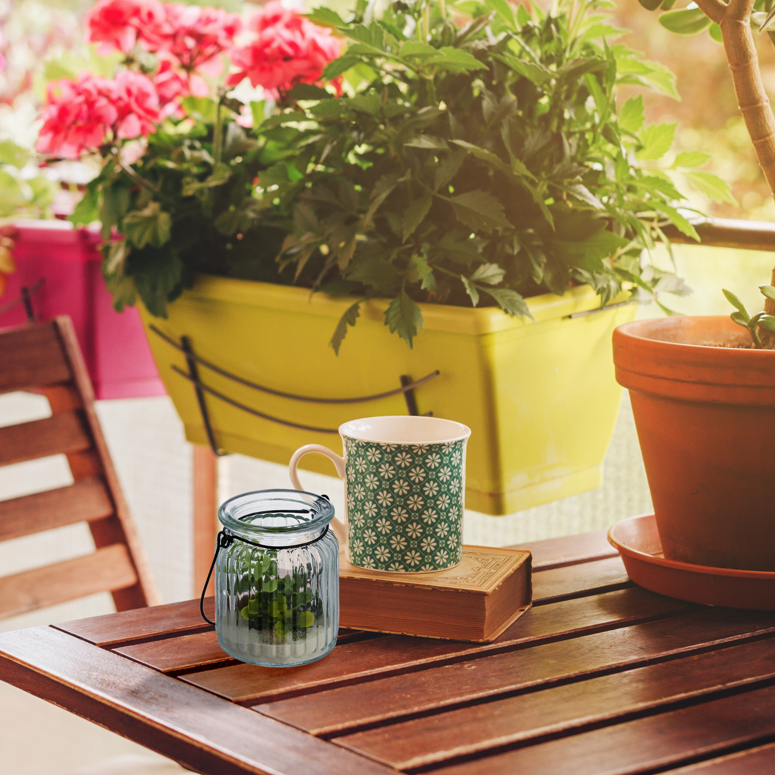 Eine Tasse, ein Buch und ein kleines Glas mit Pflanze stehen auf einem Holztisch vor Blumenkisten.