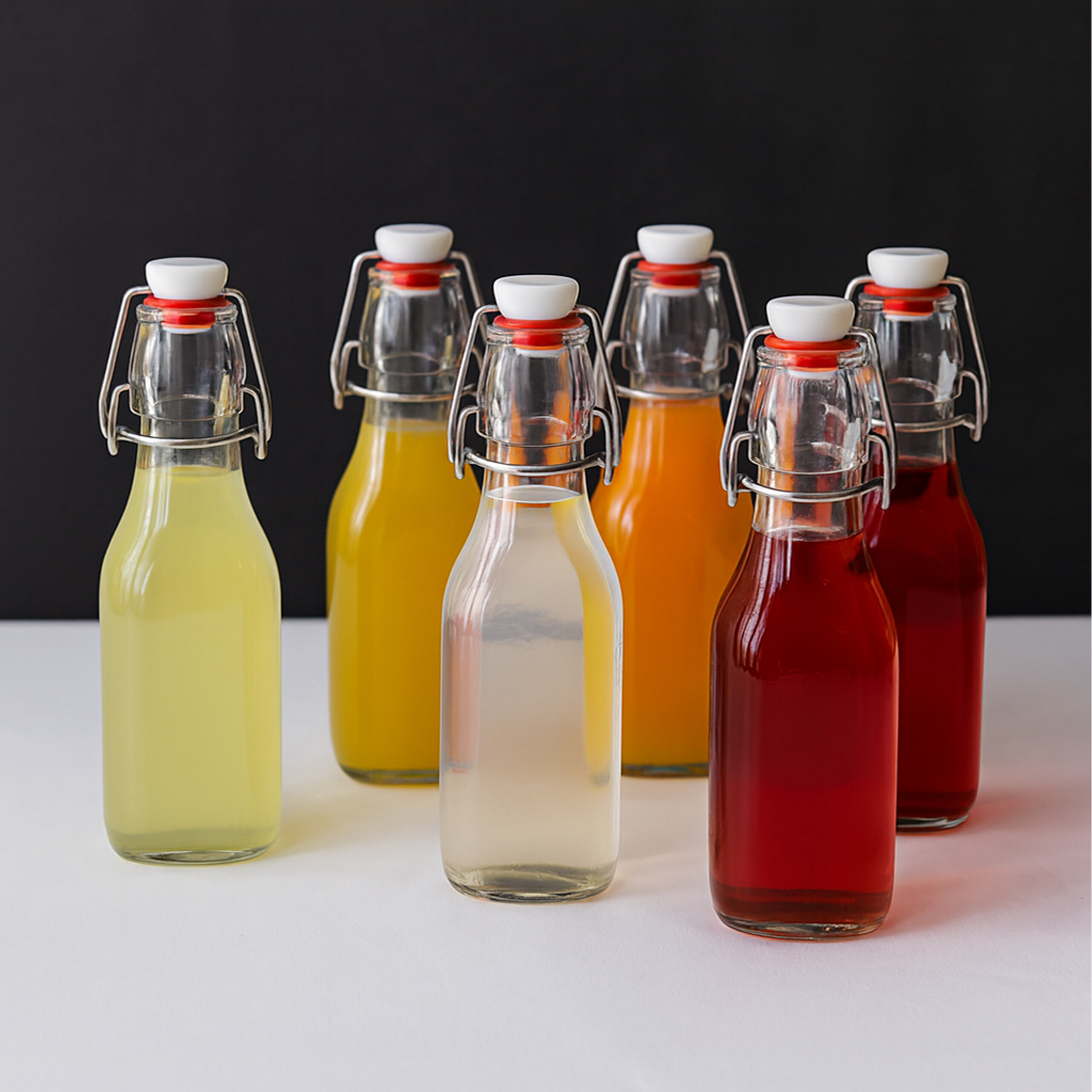Sechs Glasflaschen mit Bügelverschluss und bunten Säften stehen auf einer weißen Fläche vor dunklem Hintergrund.