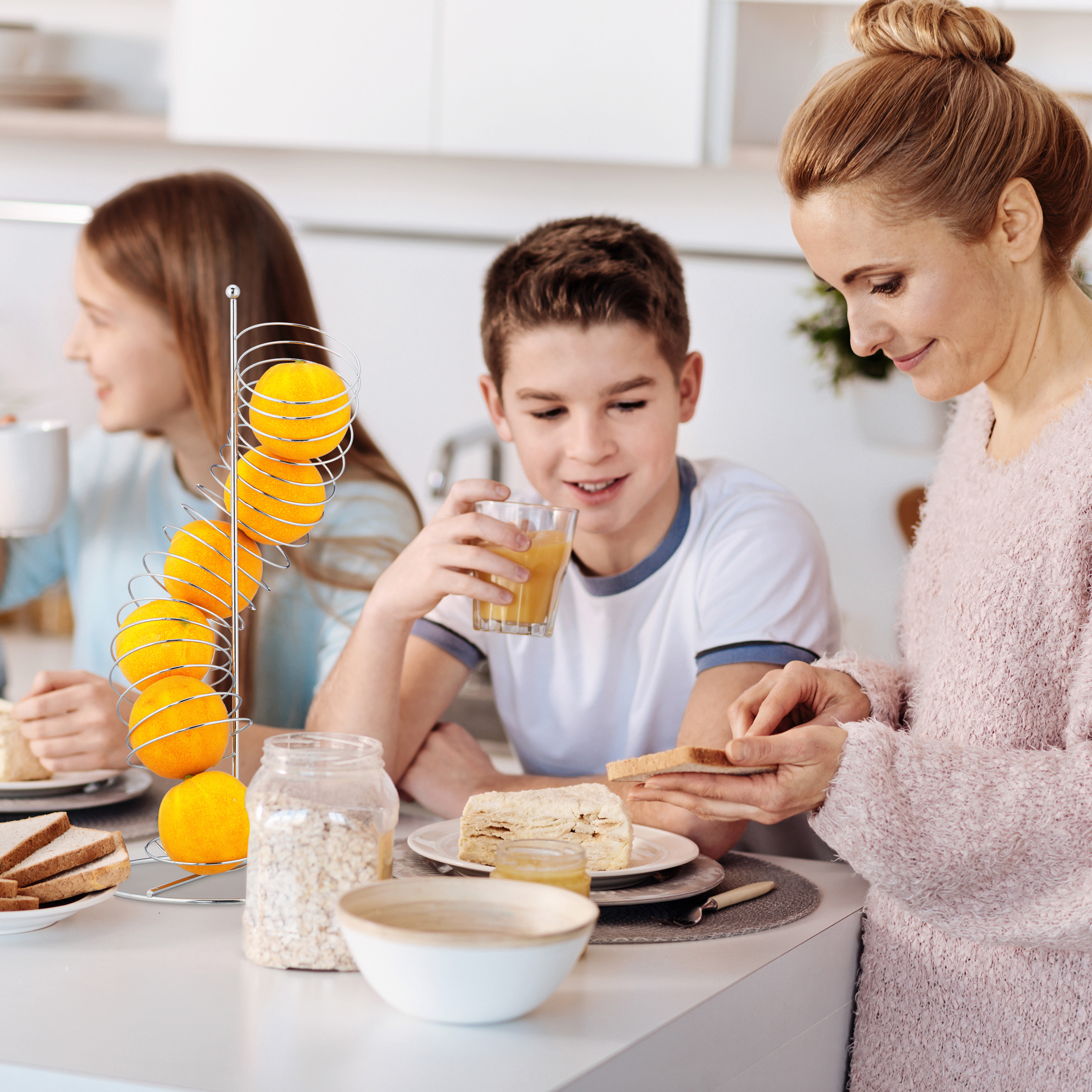 Eine Mutter bereitet in der Küche Toast zu, während ihr Sohn Saft trinkt und die Tochter im Hintergrund eine Tasse hält.