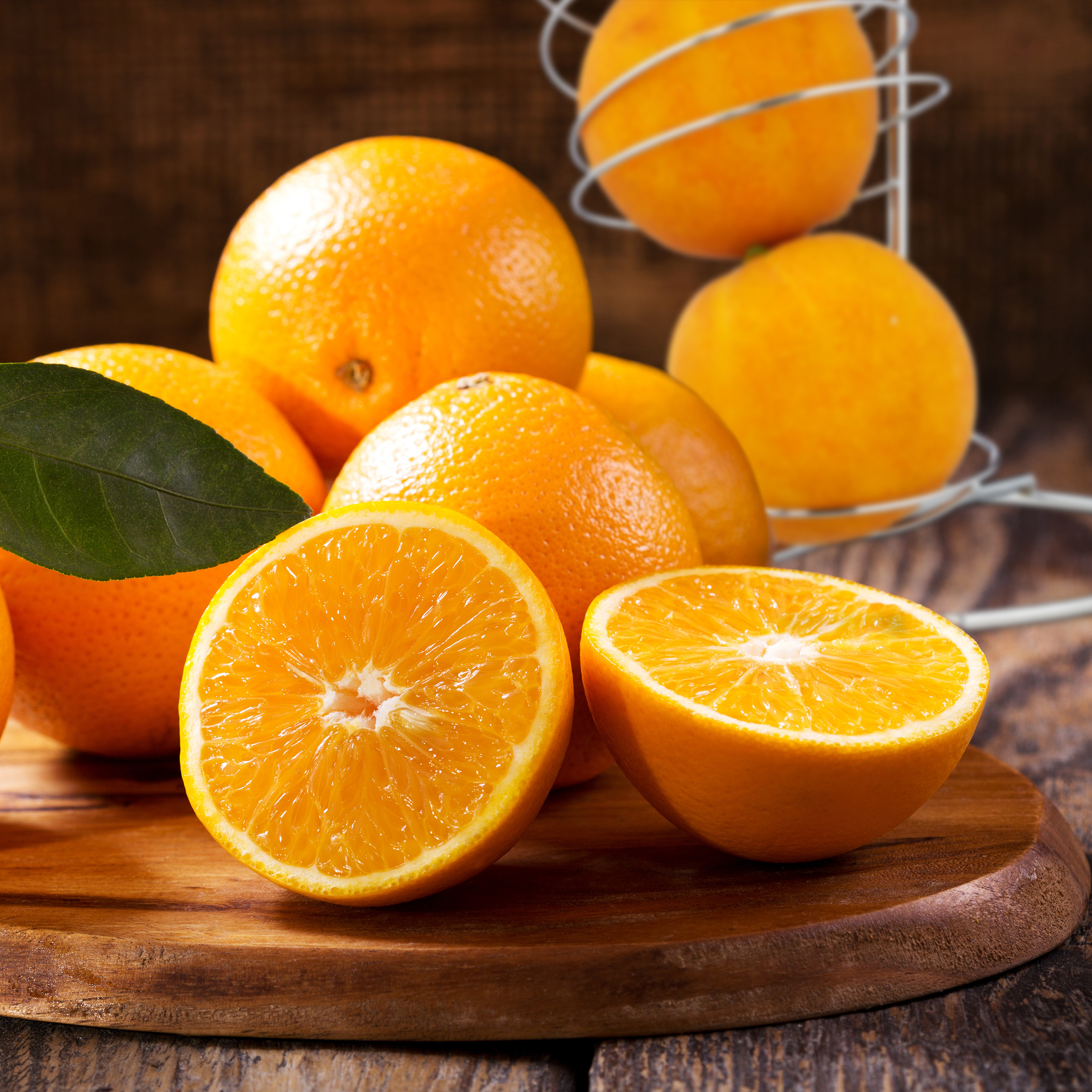 Frische Orangen, zwei davon halbiert mit einem grünen Blatt, auf einem Holzbrett vor rustikalem Holzhintergrund.