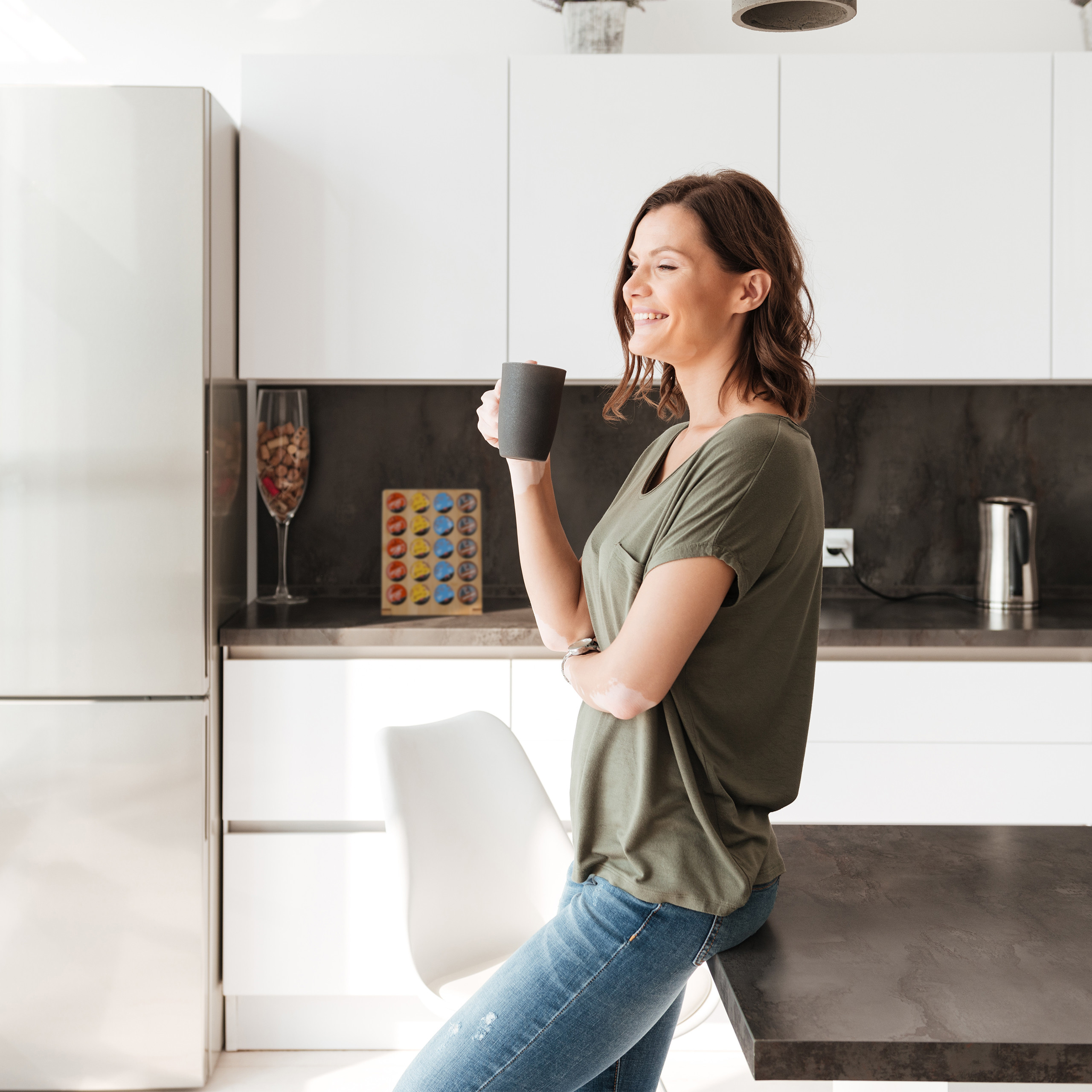 Eine lächelnde Frau steht in einer modernen Küche und hält eine graue Tasse in der Hand.