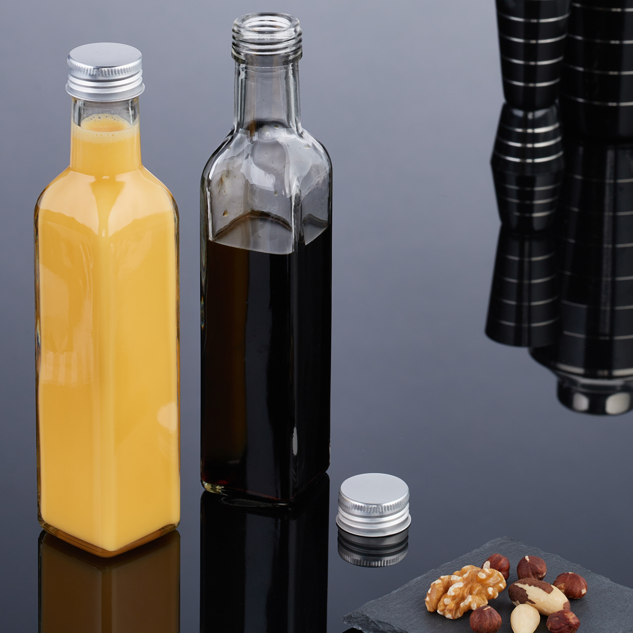 Zwei Glasflaschen mit gelber und dunkler Flüssigkeit, ein Flaschendeckel und Nüsse auf dunkler Oberfläche.
