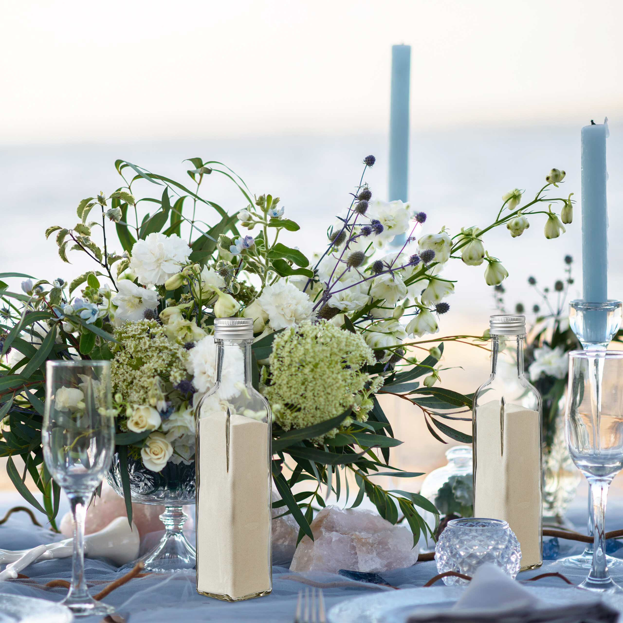 Ein Strandtisch mit weißen Blumen, Sandflaschen und blauen Kerzen.
