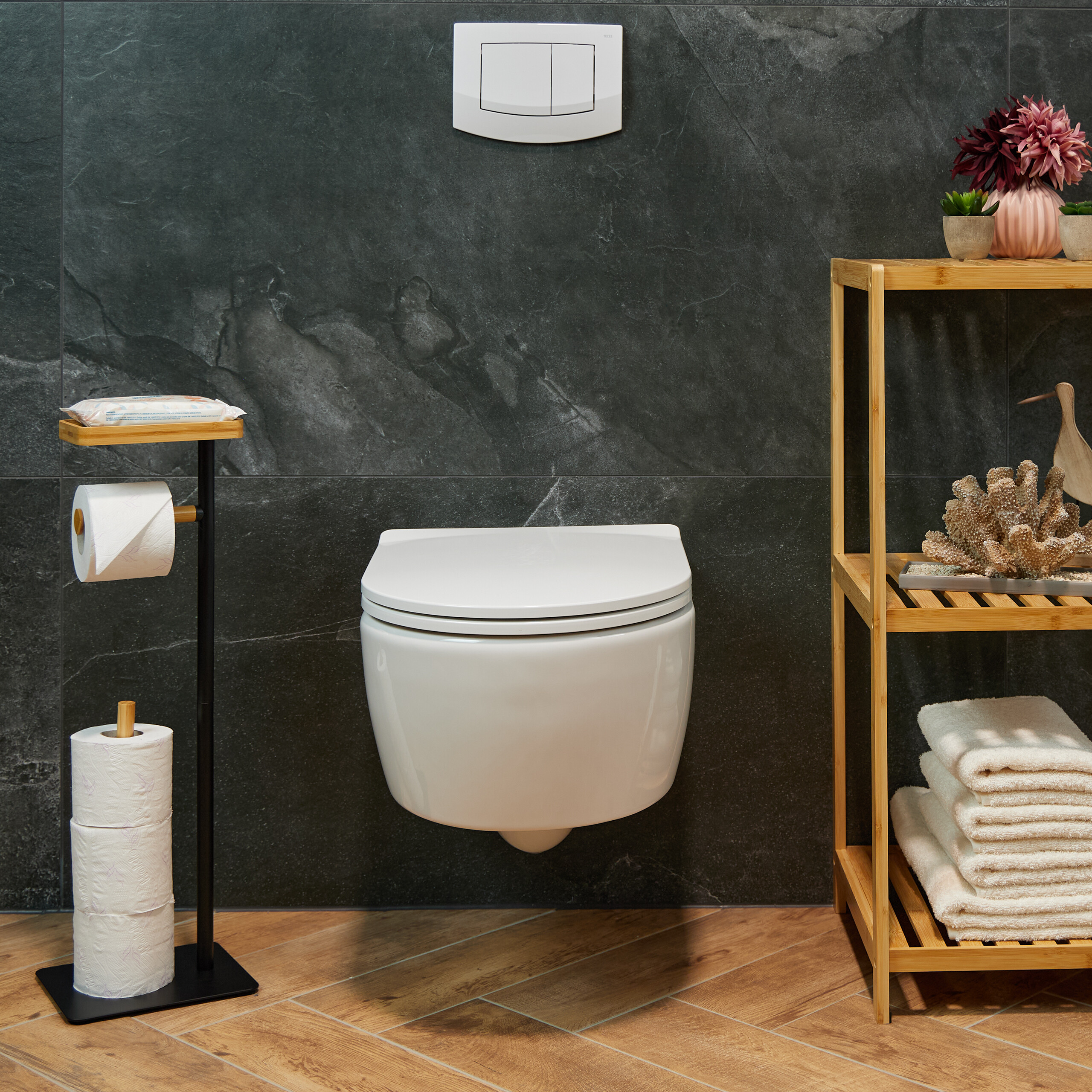 Moderne weiße Toilette vor dunklen Fliesen mit Bambusregal, Toilettenpapierhalter und Handtüchern.