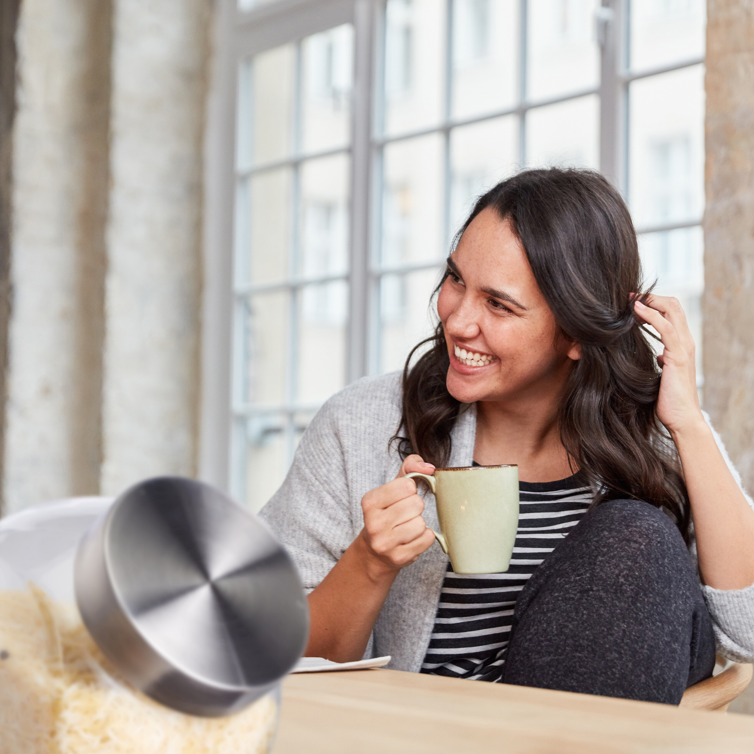 Eine lächelnde Frau sitzt am Tisch, hält eine Tasse und berührt ihr Haar vor einem großen Fenster.