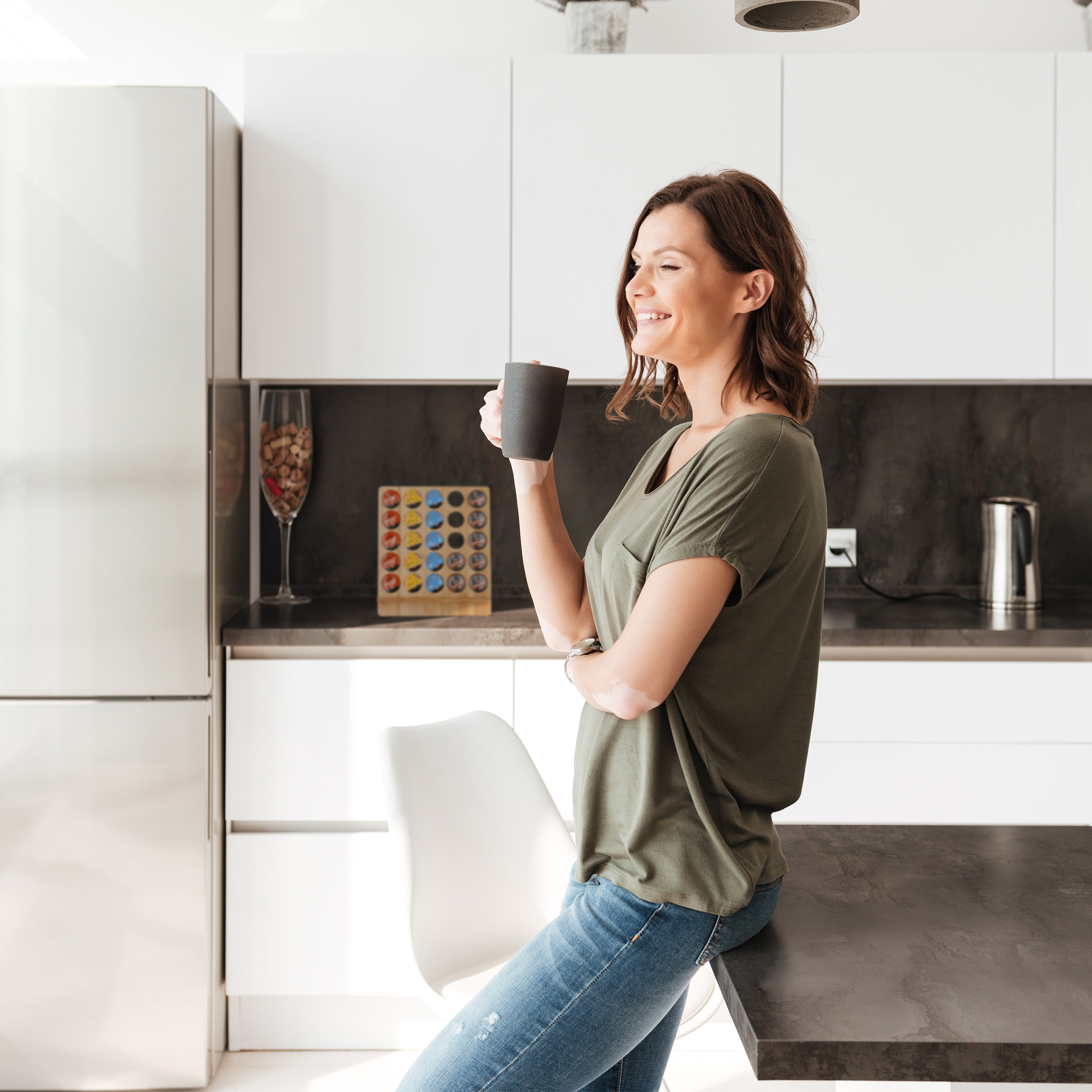 Eine Frau in grüner Bluse und Jeans lehnt an der Küchentheke und hält eine Tasse.