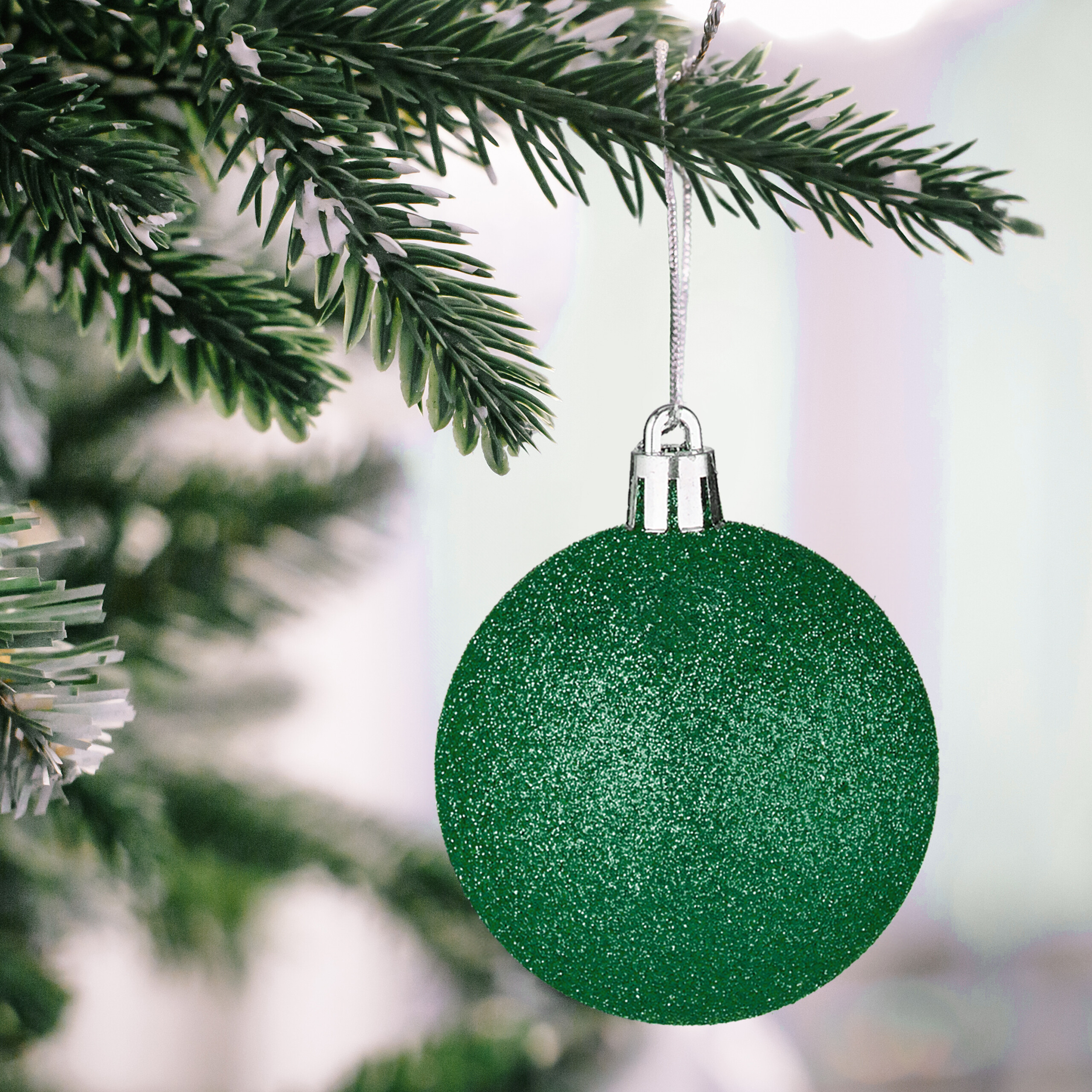 Eine glitzernde grüne Christbaumkugel hängt an einem Tannenzweig mit künstlichem Schnee.