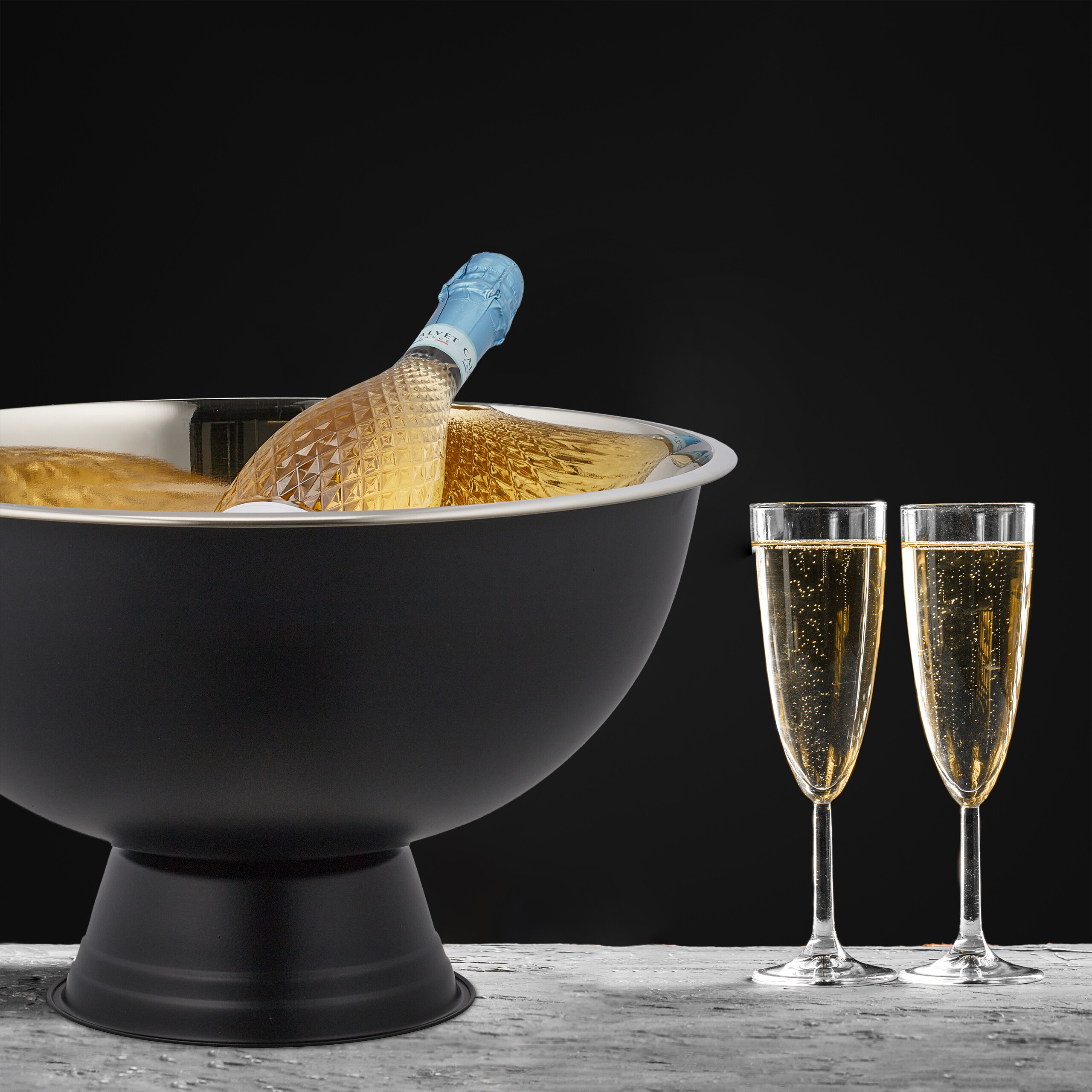 Eine Champagnerflasche im schwarzen Kühler mit zwei Gläsern auf grauem Tisch vor schwarzem Hintergrund.