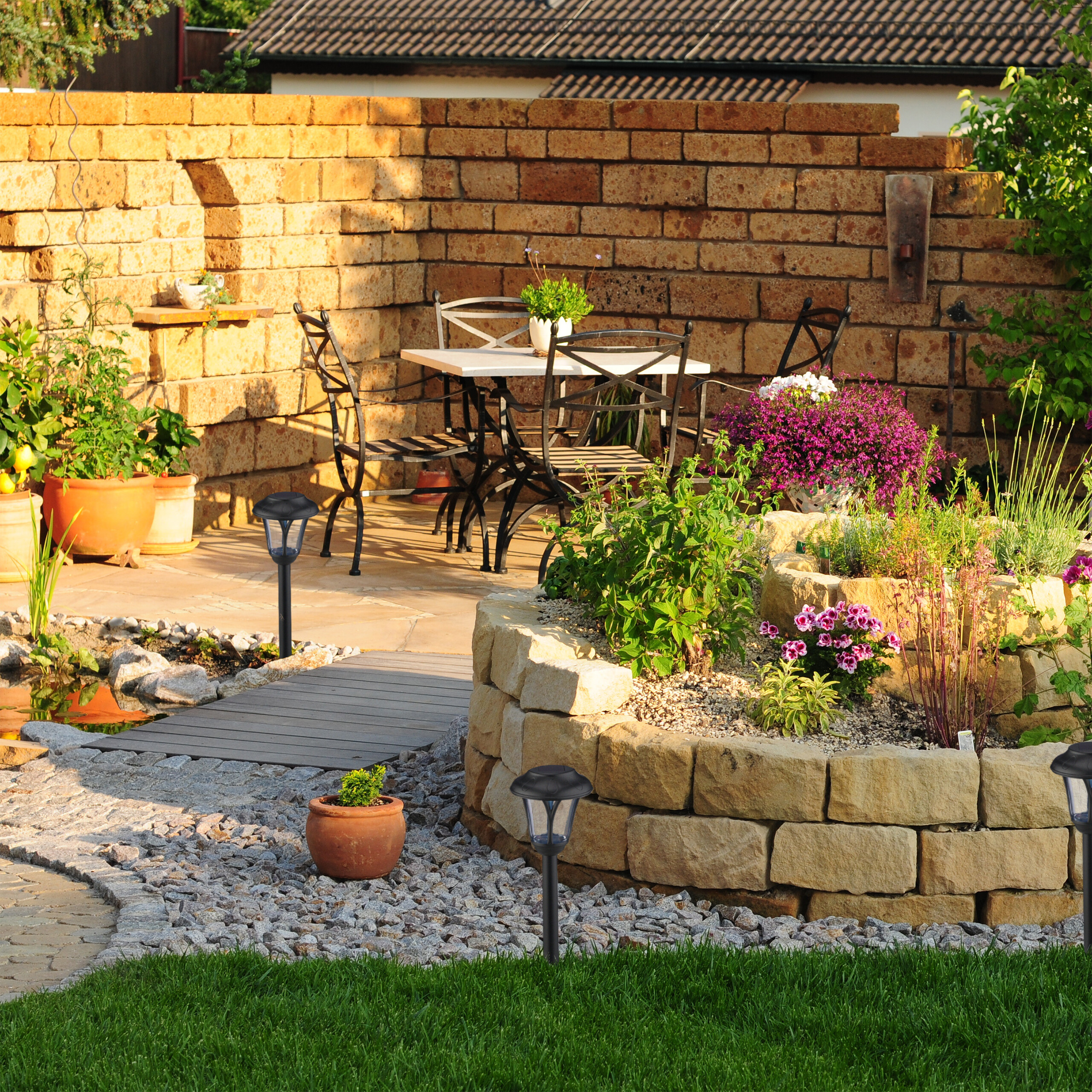 Ein sonniger Garten mit Backsteinmauer, Steinbeet, Holzbrücke, Teich und Metalltischset.