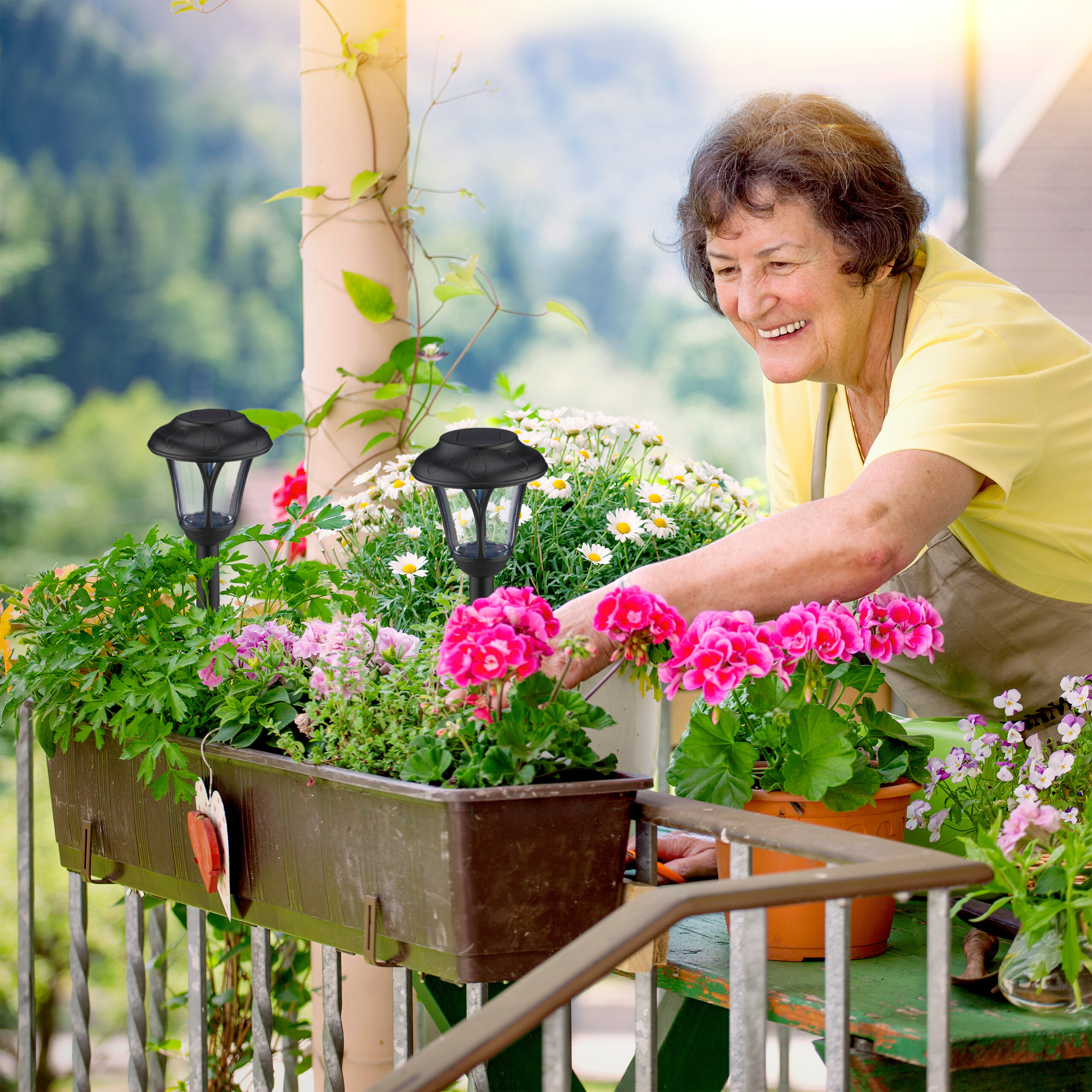 Lchelnde ltere Frau pflegt Blumenksten und Topfpflanzen auf einem sonnigen Balkon mit zwei schwarzen Solarleuchten.
