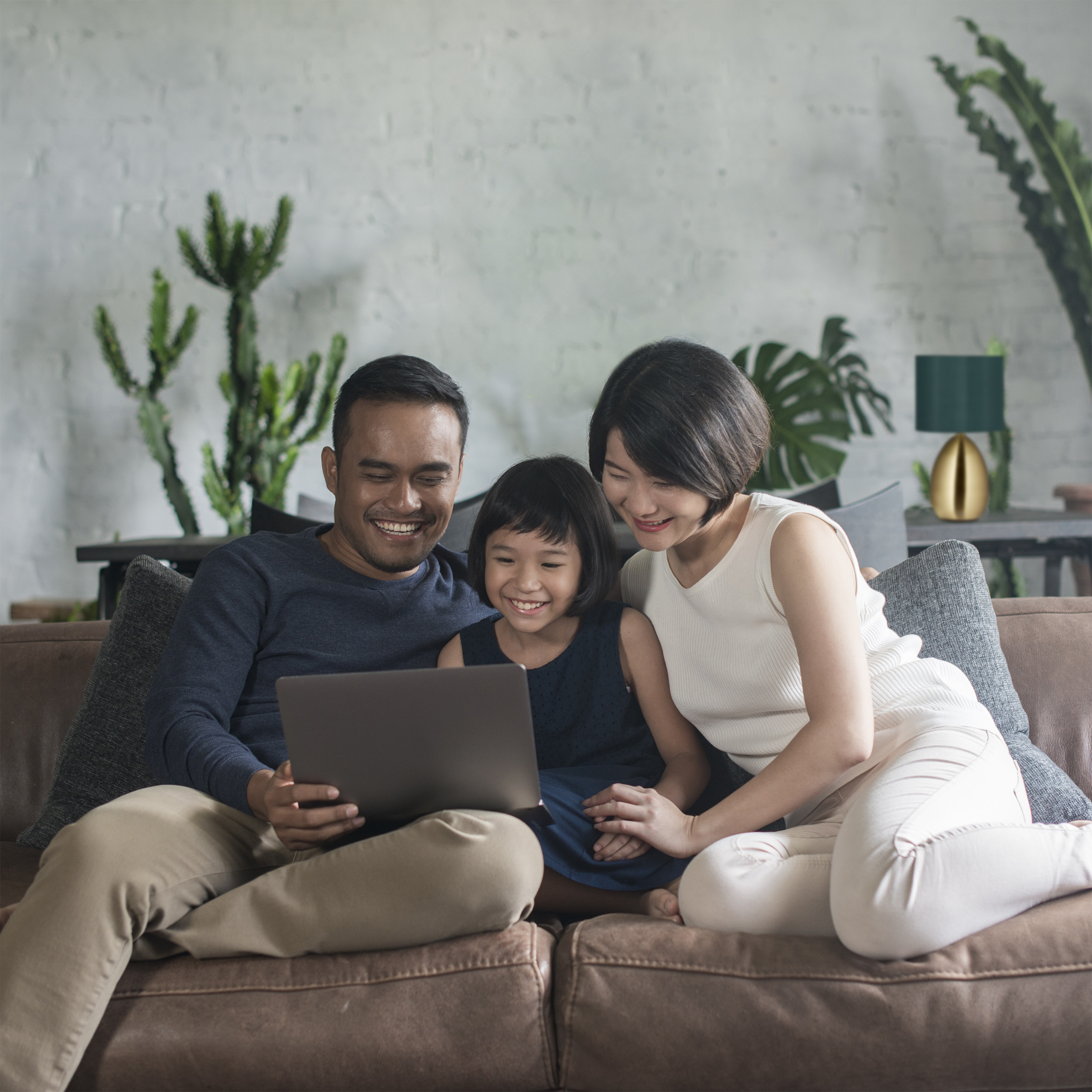 Eine glückliche asiatische dreiköpfige Familie sitzt auf einem braunen Sofa und schaut in einem hellen Zimmer auf einen Laptop.