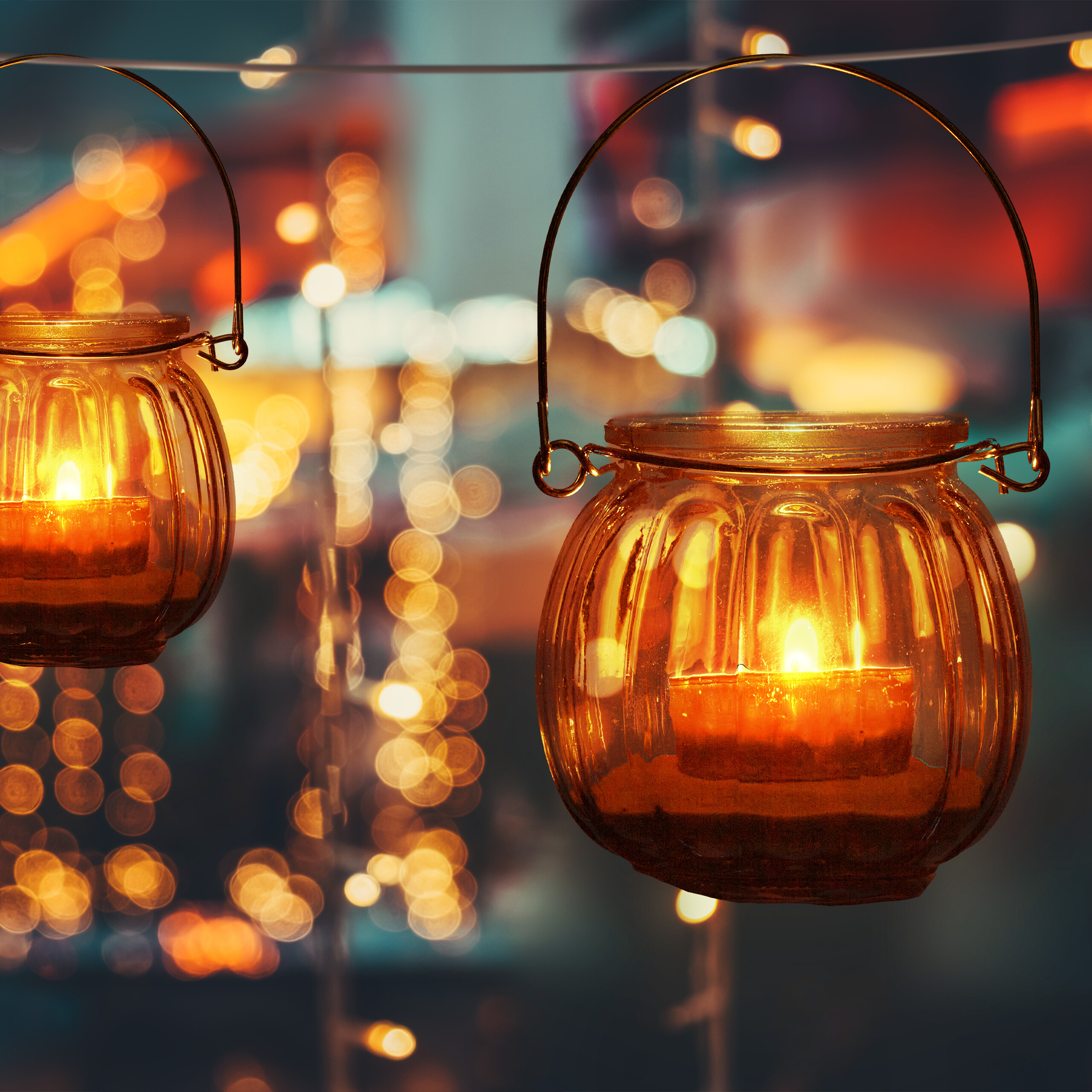 Zwei bernsteinfarbene Glasteelichter mit brennenden Kerzen hängen draußen vor einem verschwommenen Bokeh-Hintergrund.