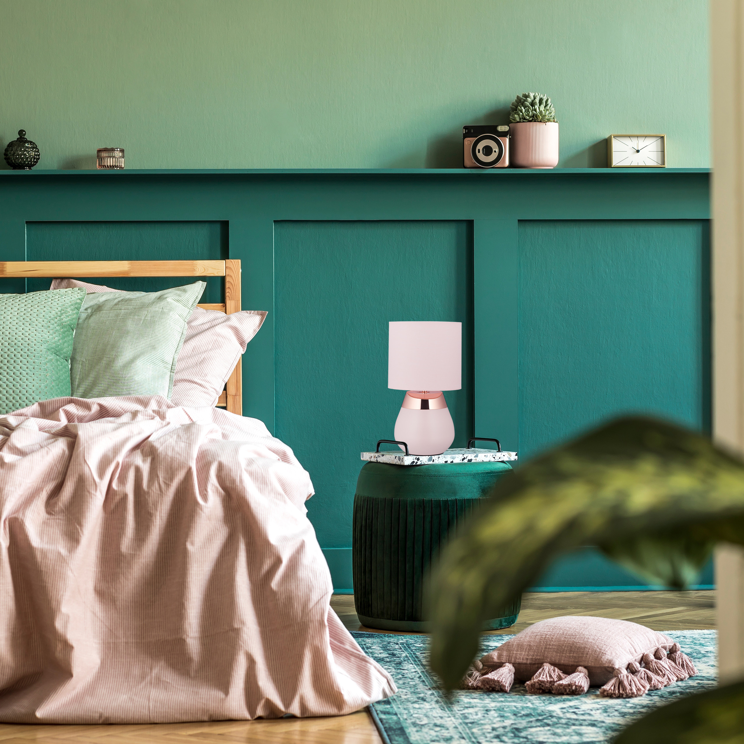 Ein Schlafzimmer mit grünen Wänden, einer rosa Lampe auf einem grünen Hocker und einem Bett mit rosa und grünen Kissen.