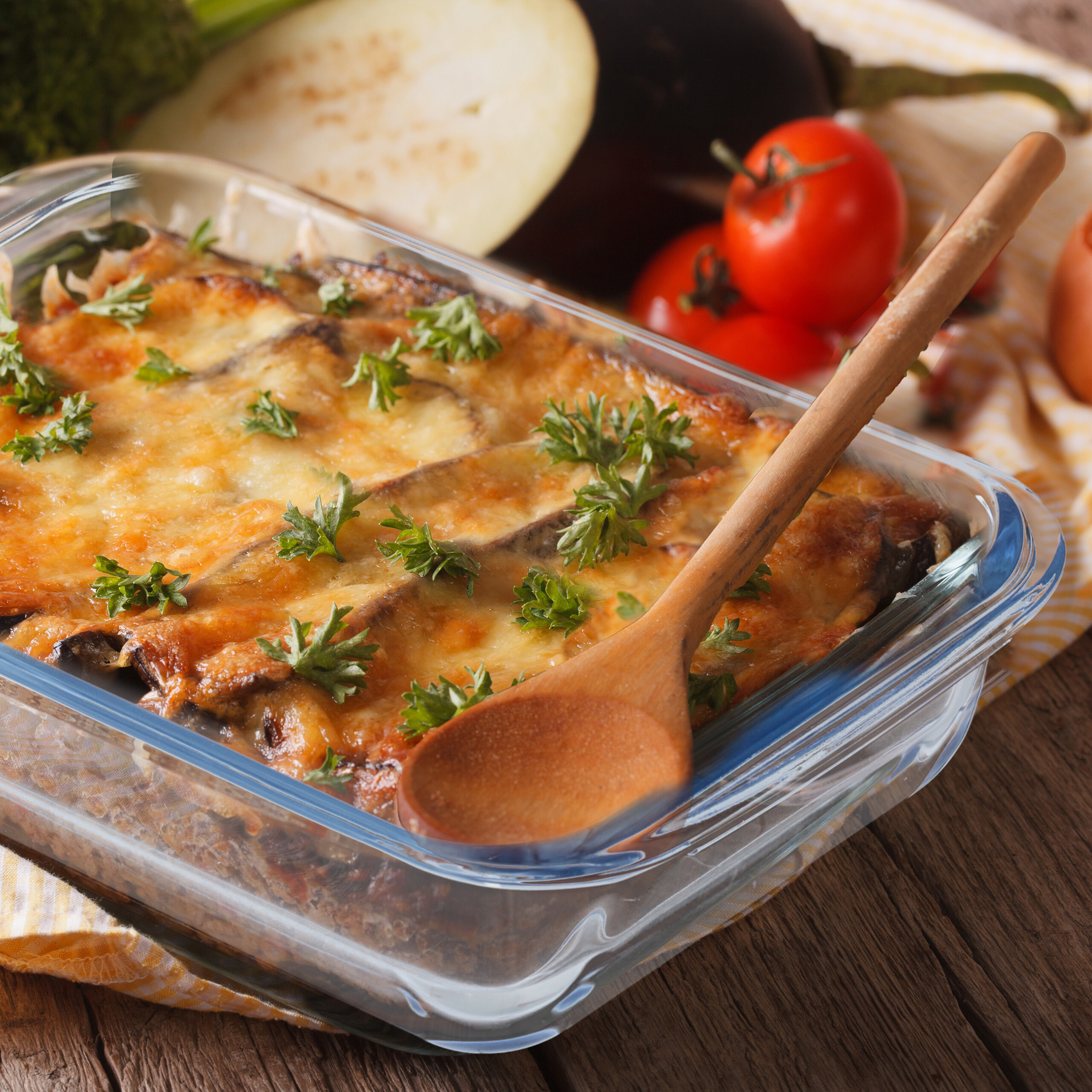 Frisch gebackener Auberginen-Auflauf mit geschmolzenem Käse und Kräutern in einer Glasform, begleitet von frischem Gemüse.