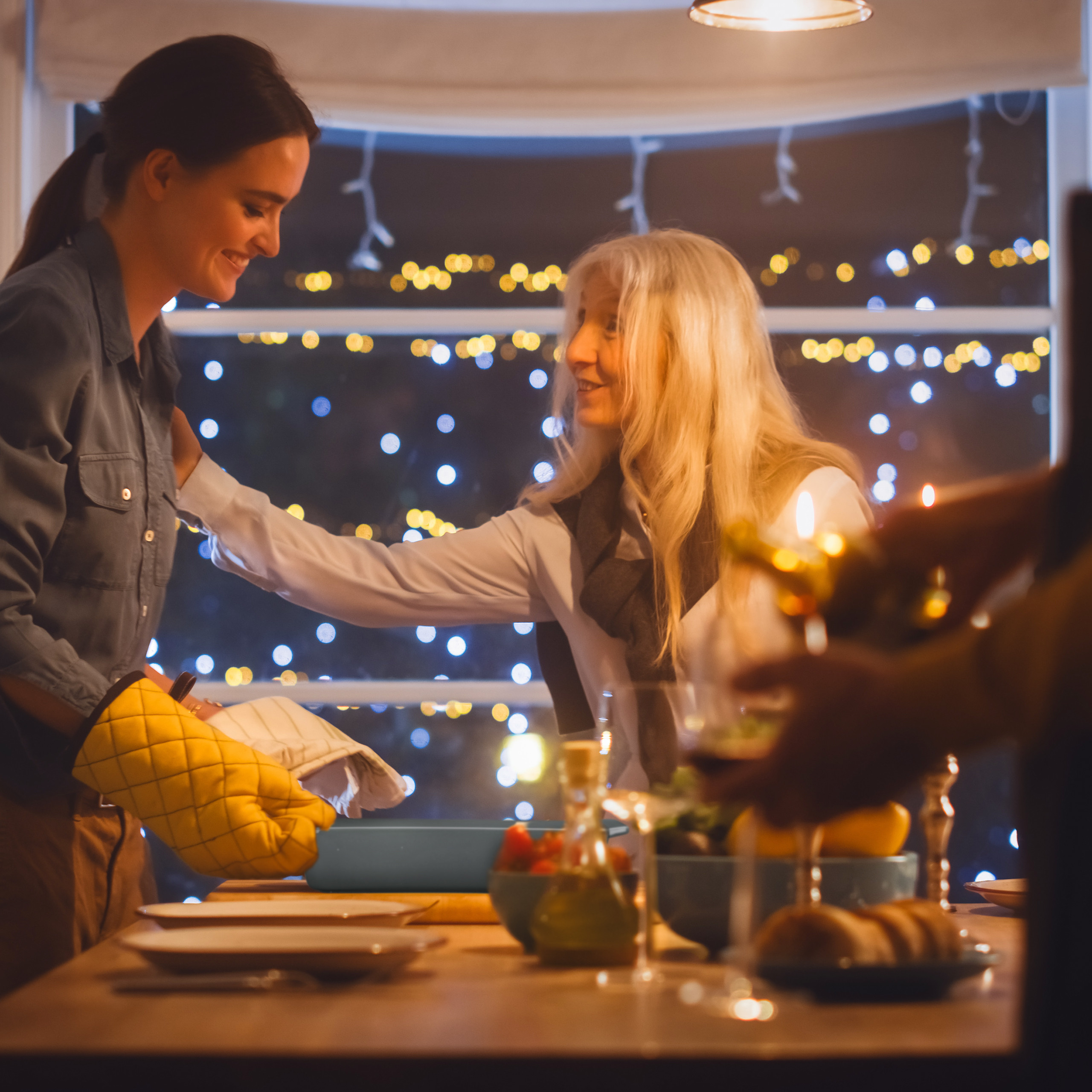 Zwei Frauen lächeln beim Abendessen in einer gemütlichen Küche mit verschwommenen Lichtern.