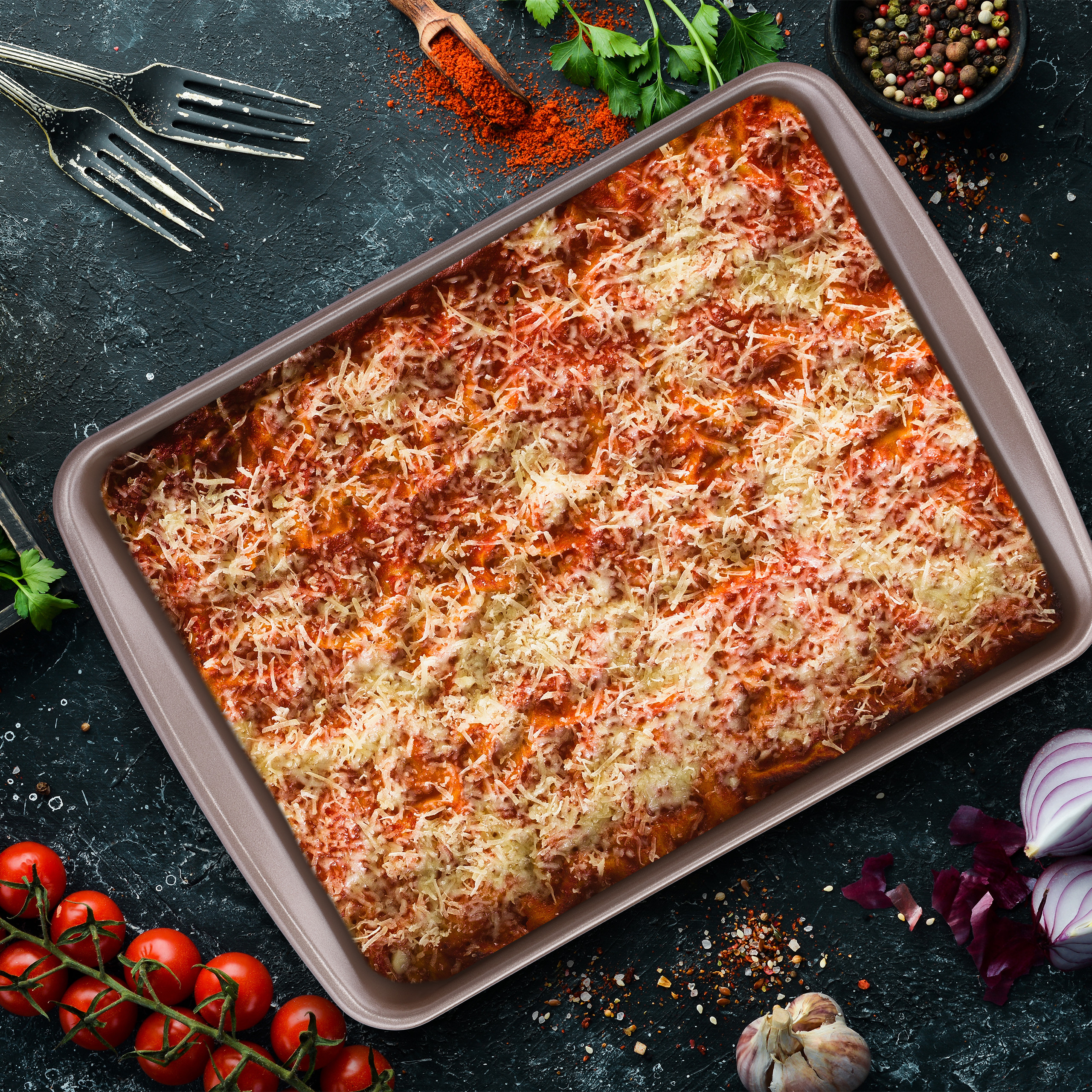 Frisch gebackene Lasagne in einer Backform auf dunklem Tisch mit Tomaten und Gewürzen.
