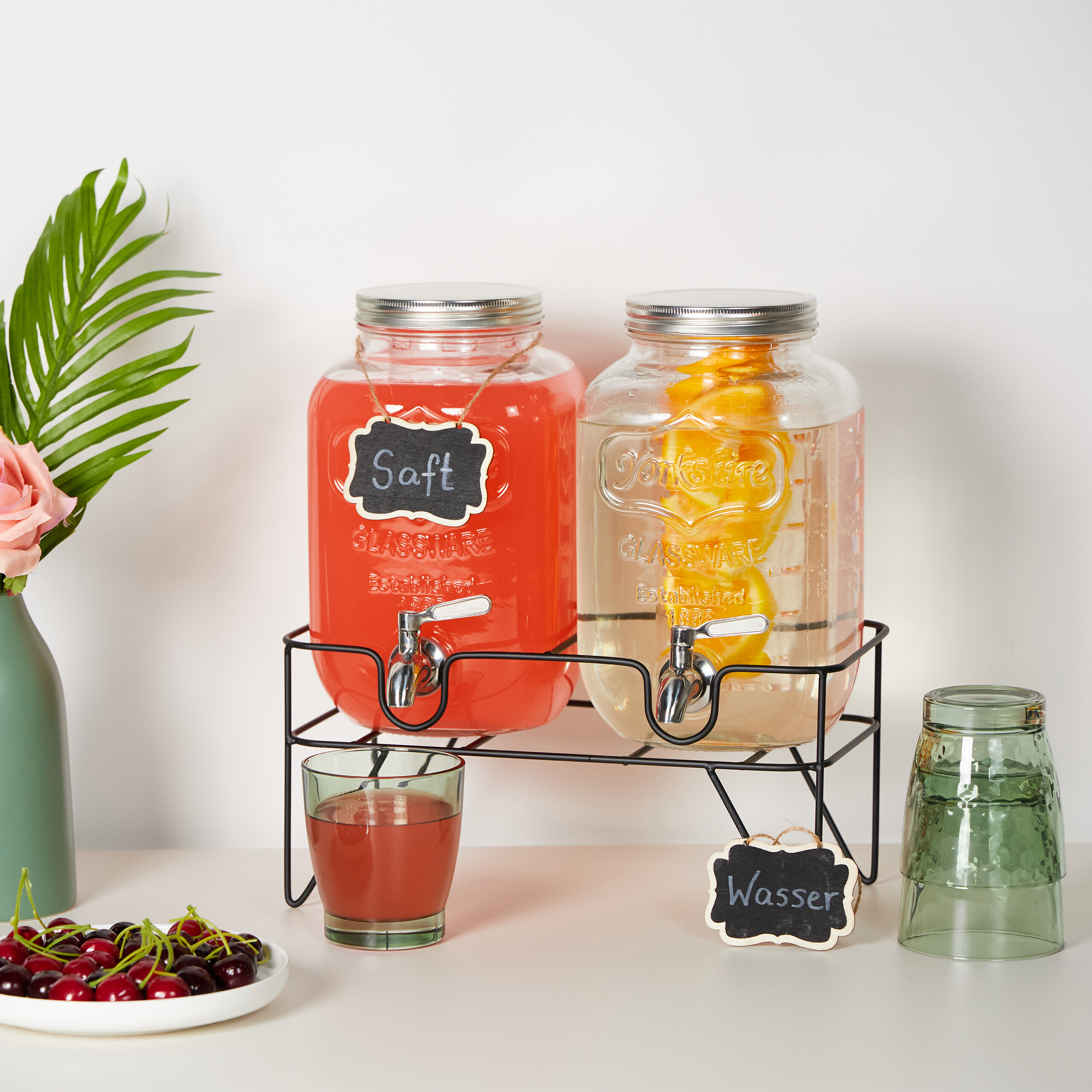 Zwei Getränkespender mit Saft und Wasser auf einem Ständer, daneben Kirschen und Gläser.